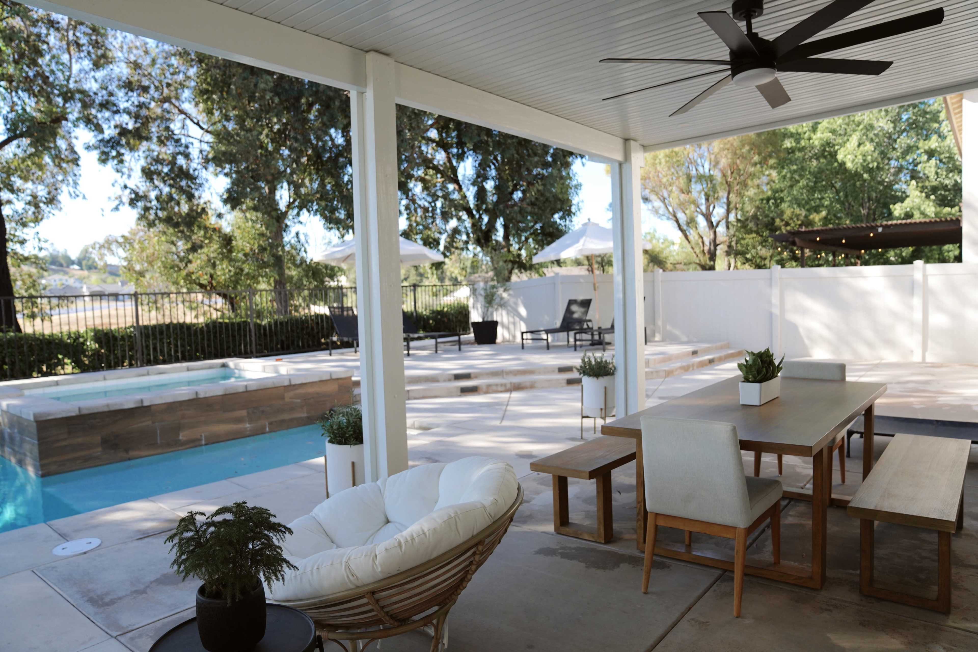 A covered patio features a dining table with benches and a lounge chair, overlooking a swimming pool and landscaped area.