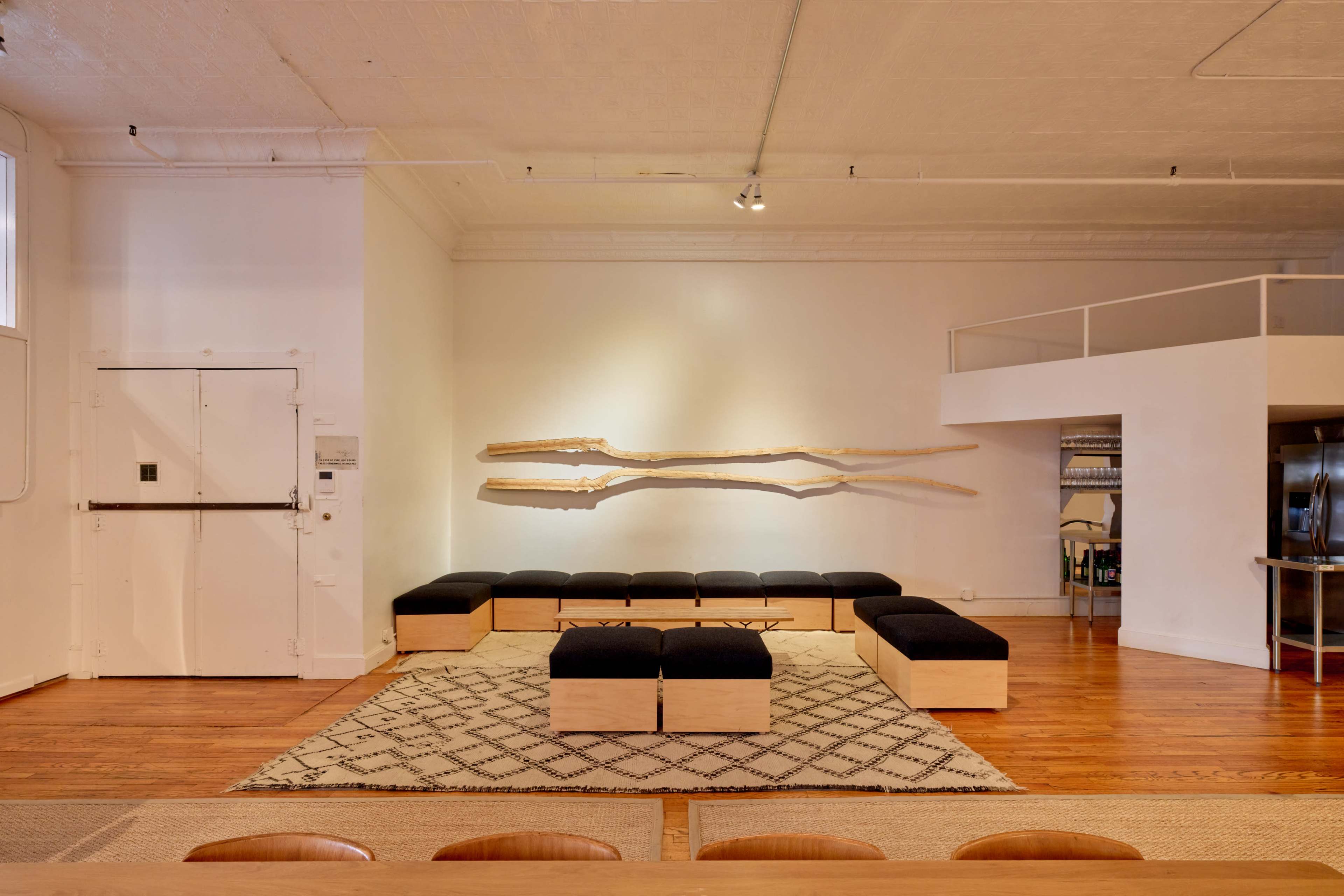 The image shows a spacious interior with a wooden floor, minimalistic seating area featuring long benches, and an artistic wooden wall piece.