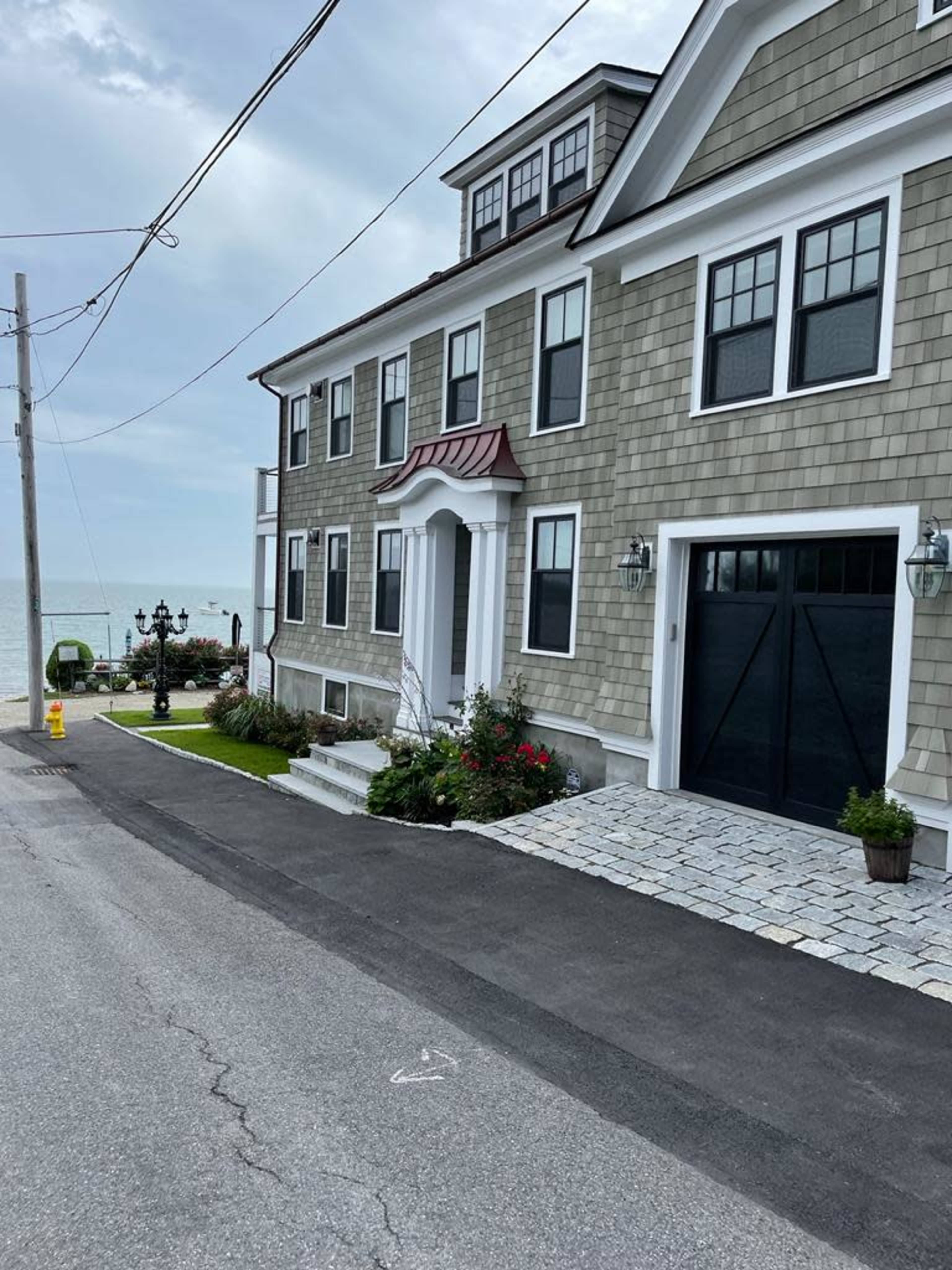 A large, two-story green shingle house with white trim and a garage is located near a beach, with a paved driveway and decorative landscaping.