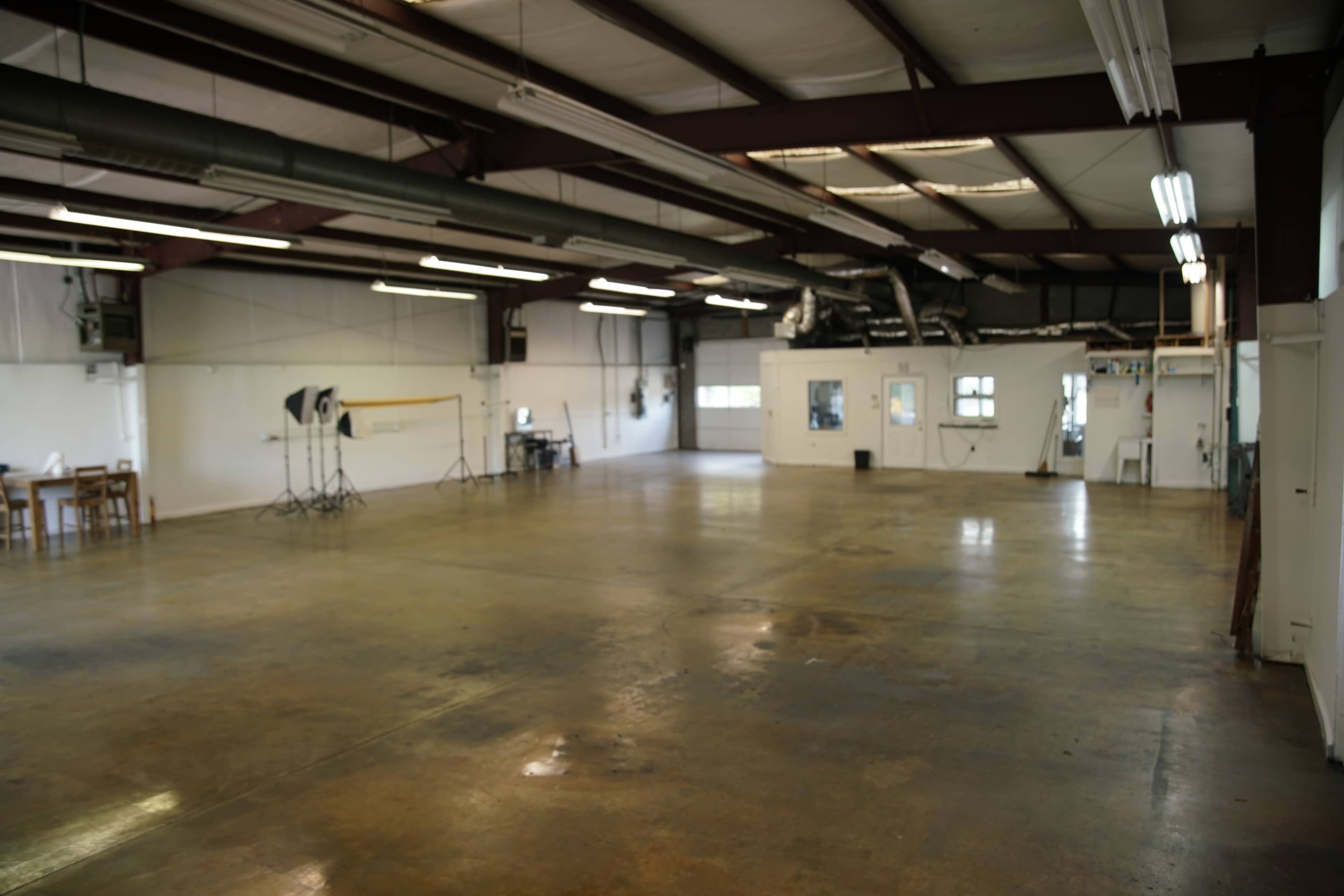 The image shows an empty industrial space with concrete flooring, large windows, and equipment for photography set up in one corner.