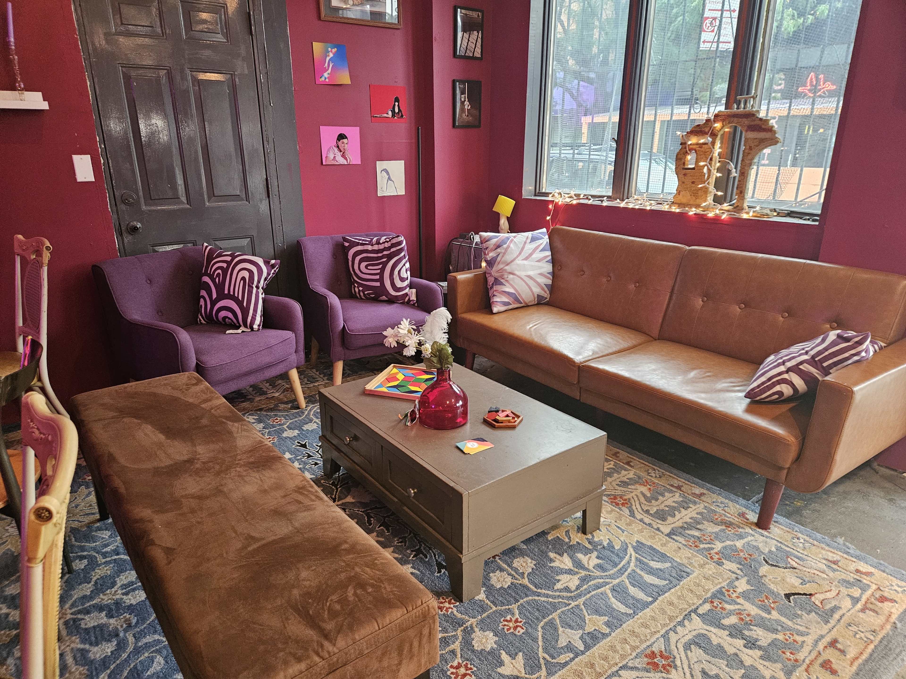 The image shows a cozy living room with purple and brown sofas, a wooden coffee table, and colorful wall art against a deep red backdrop.