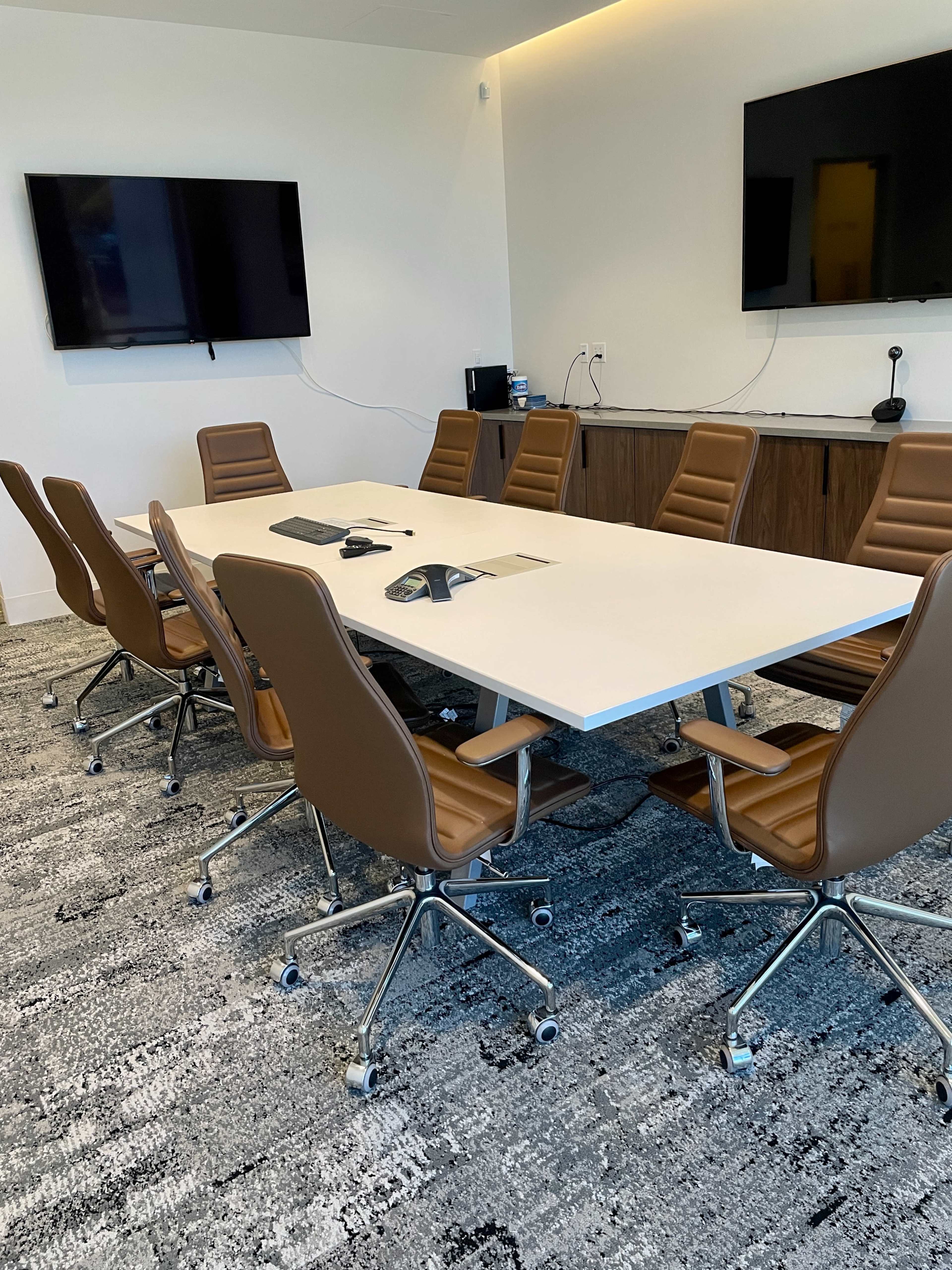 A modern conference room with a long white table surrounded by brown upholstered chairs, two wall-mounted screens, and a carpeted floor.