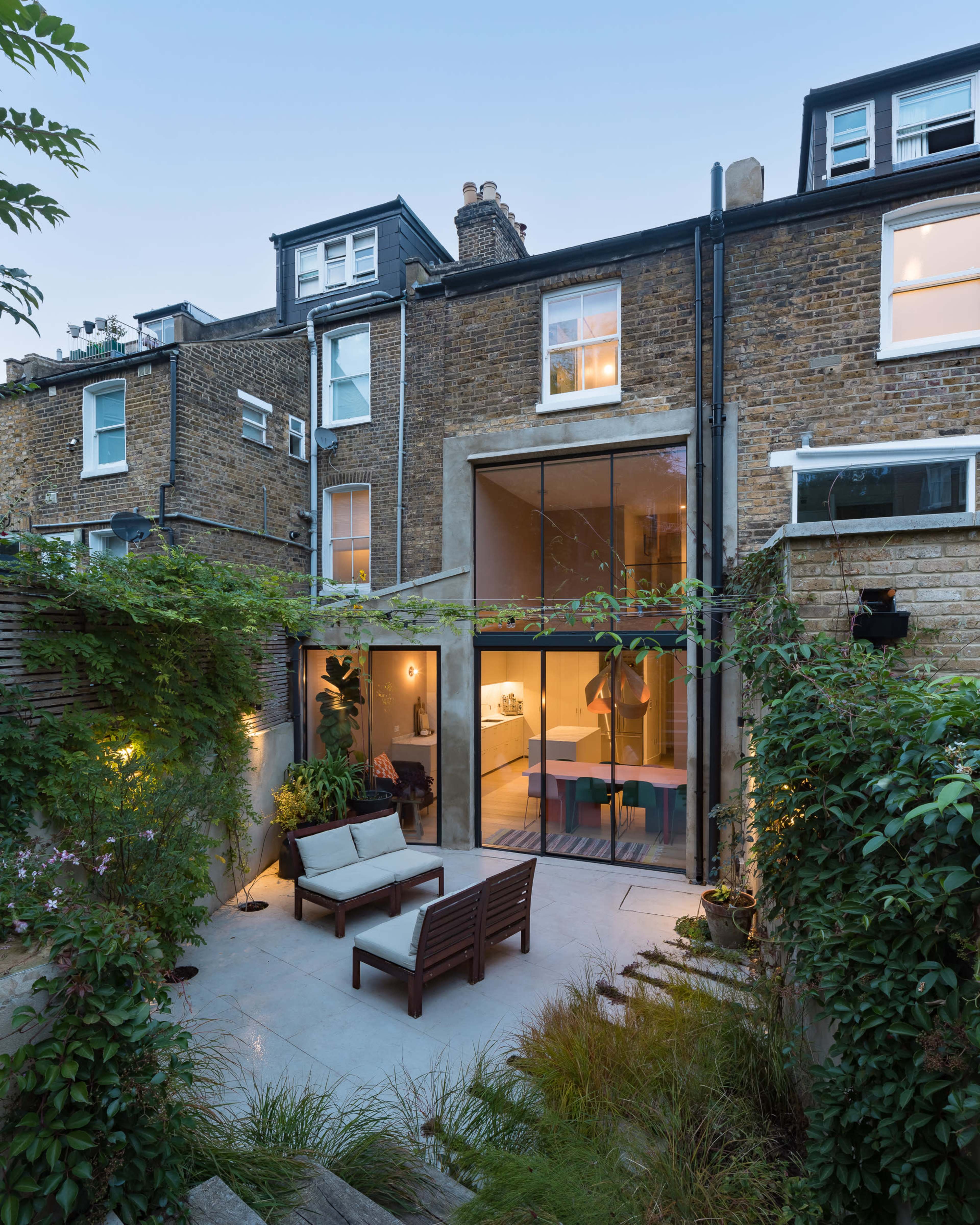 A modern two-story brick house with large glass doors opens into a backyard patio featuring seating and lush greenery.
