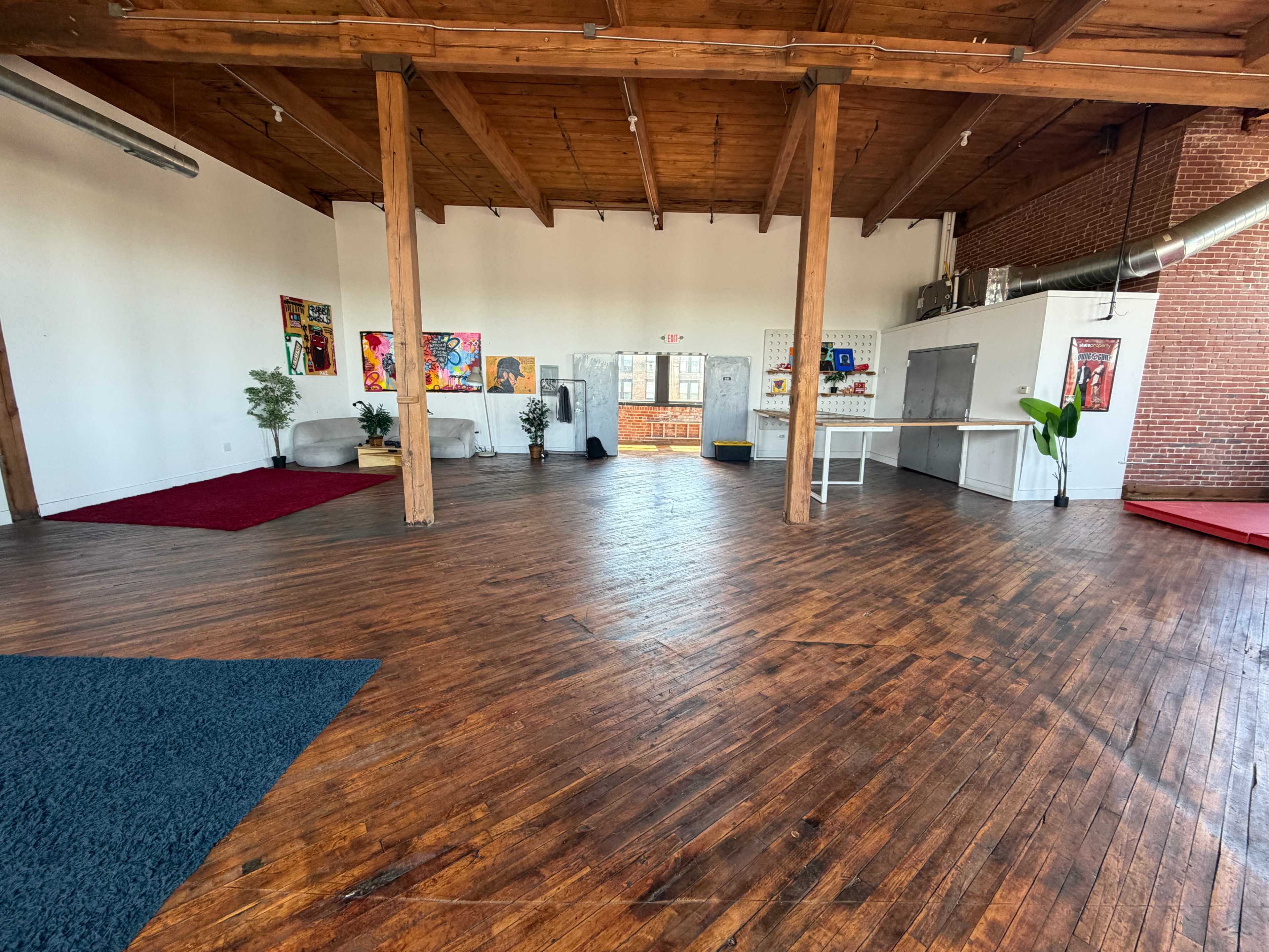 The image shows a spacious, empty room with wooden floors, wooden beams, and various artworks on the walls.