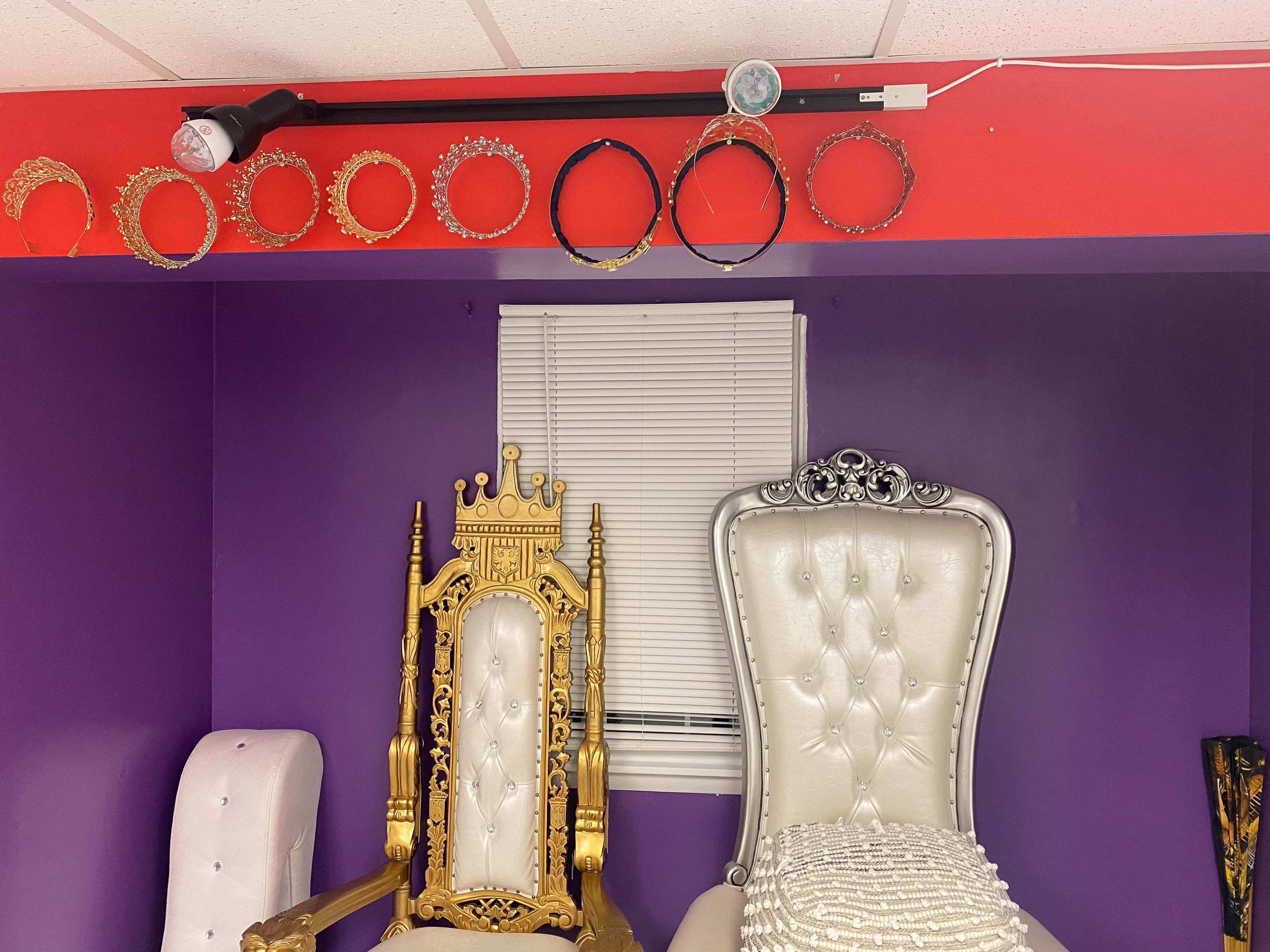 Two decorative chairs, one gold and ornate and the other silver with a tufted design, positioned against a purple wall, with a line of crowns displayed above them.