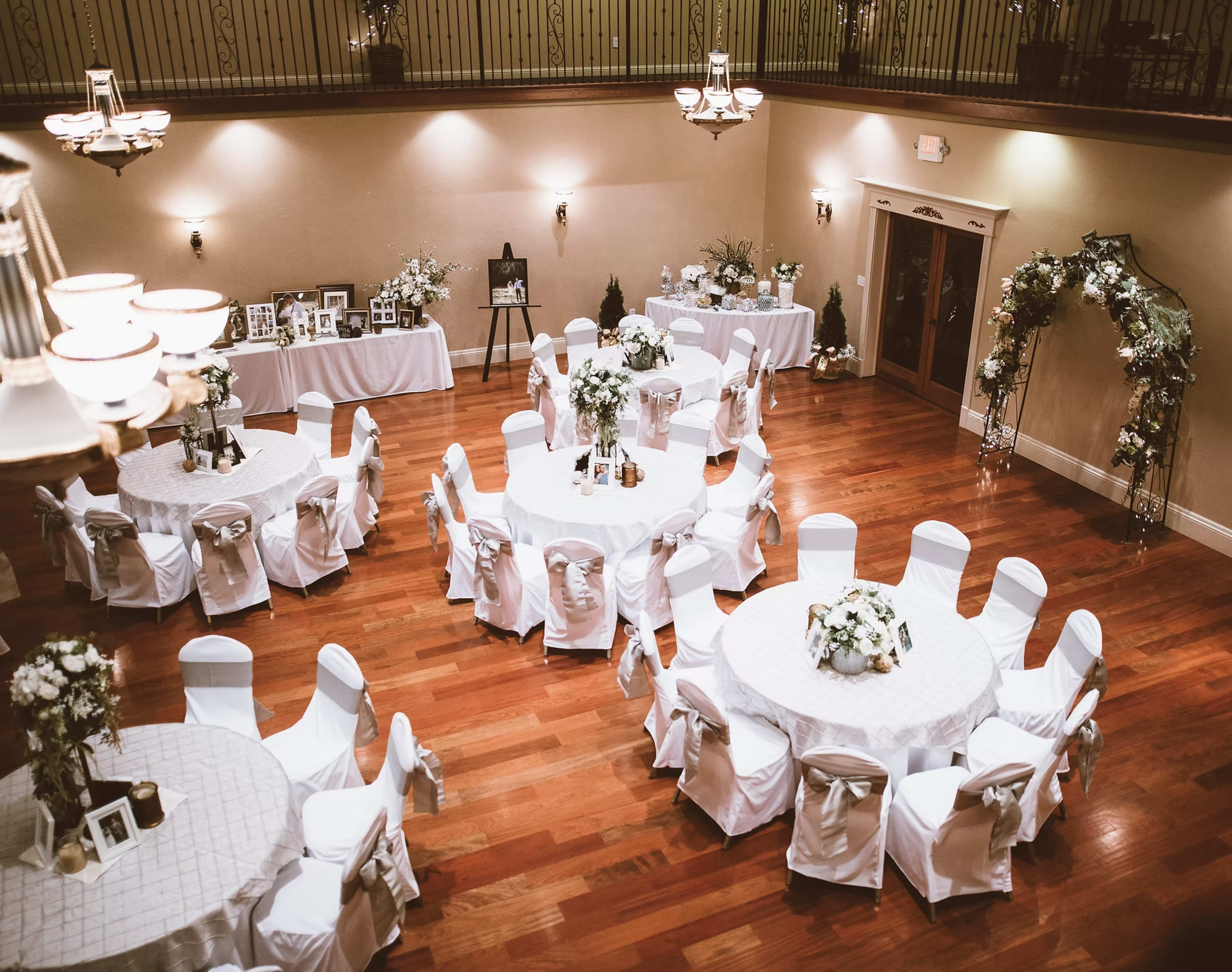 An elegantly arranged event space features round tables draped in white cloth, decorated with floral centerpieces, and surrounded by white chairs, set against a backdrop of wooden flooring and soft lighting.