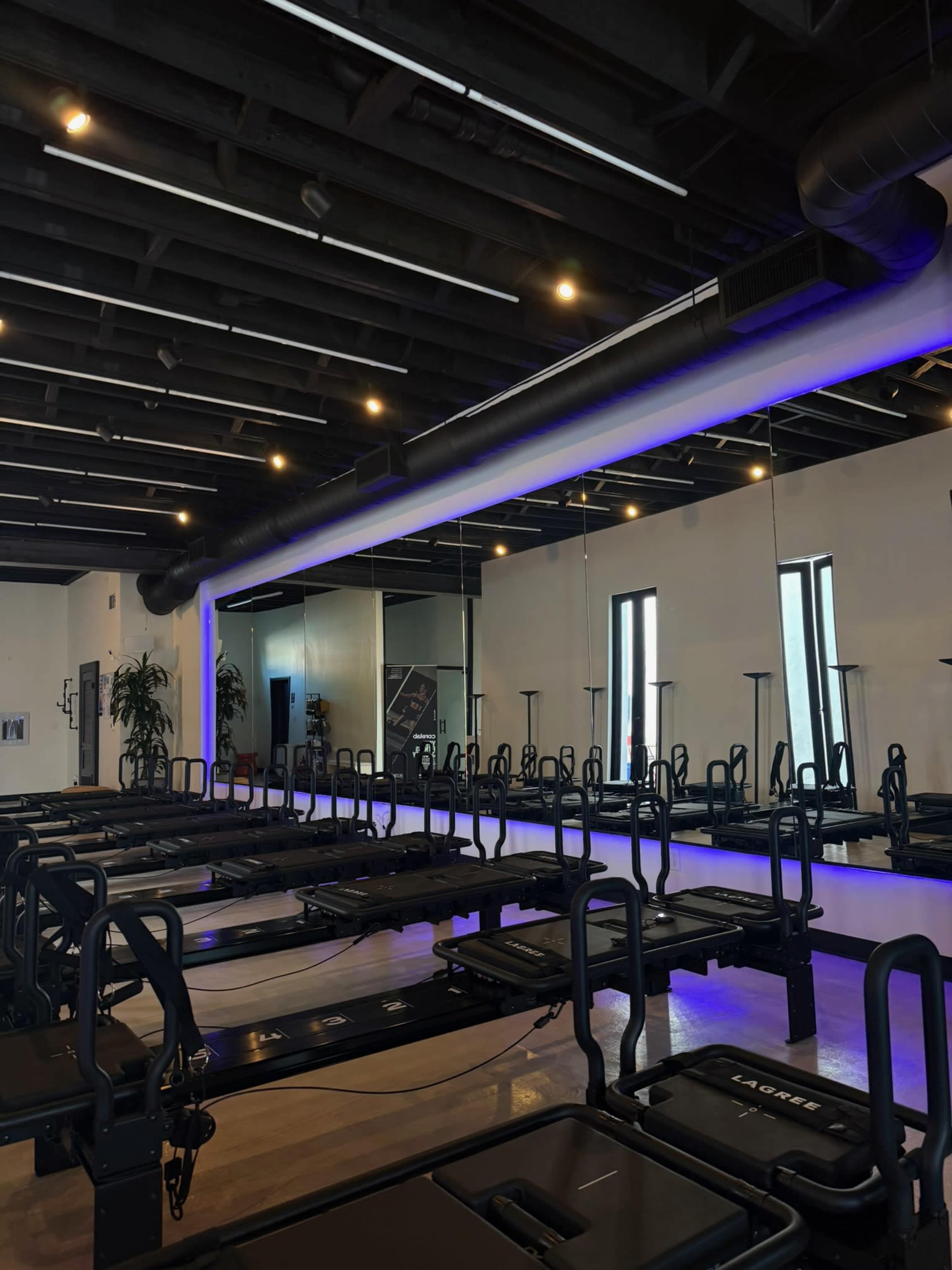 The image shows a modern fitness studio with multiple reformer machines arranged in rows, under a ceiling with exposed beams and bright overhead lighting.