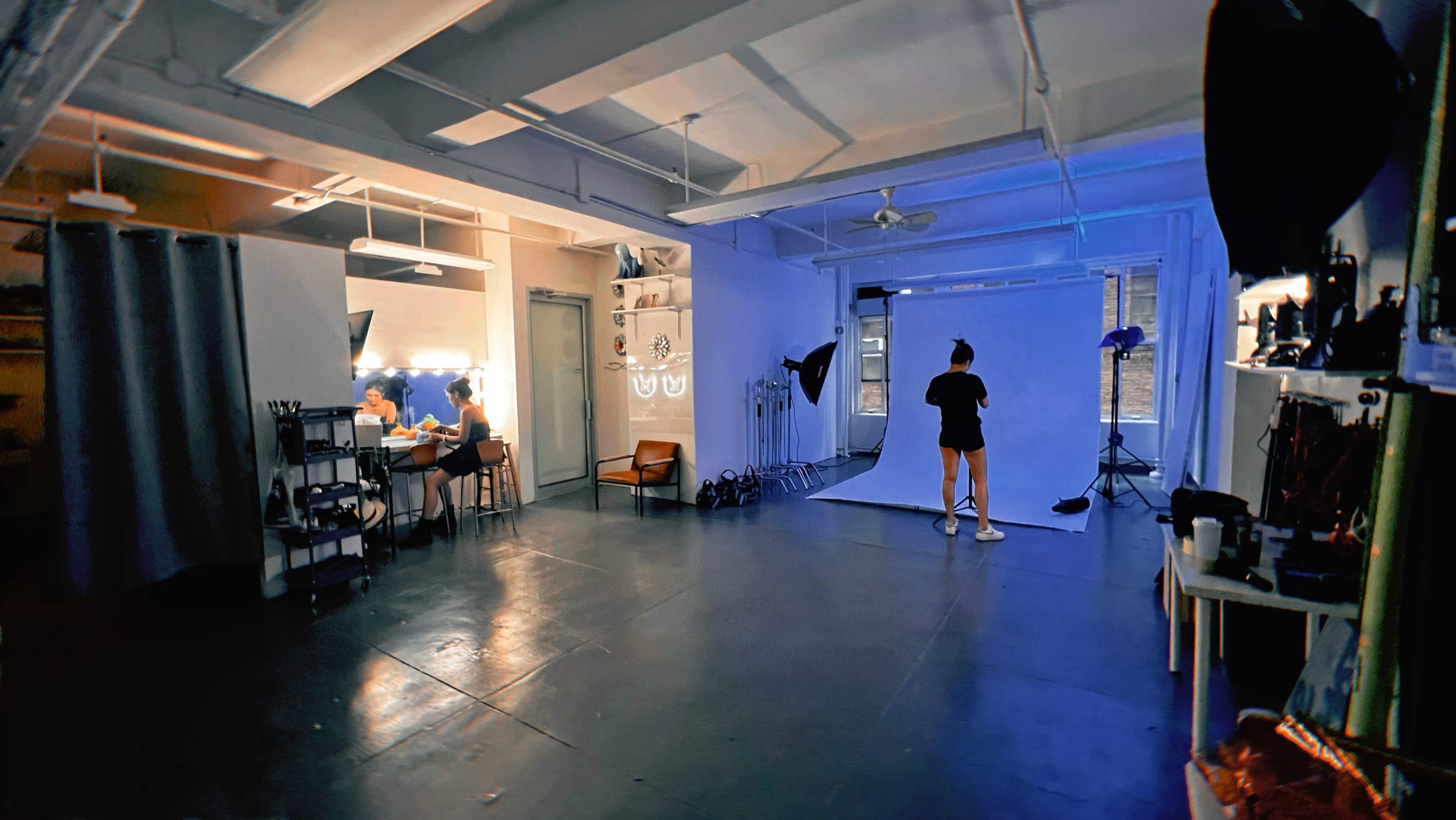 A spacious studio with a blue backdrop, equipped with lighting equipment, and a table where two people are seated.