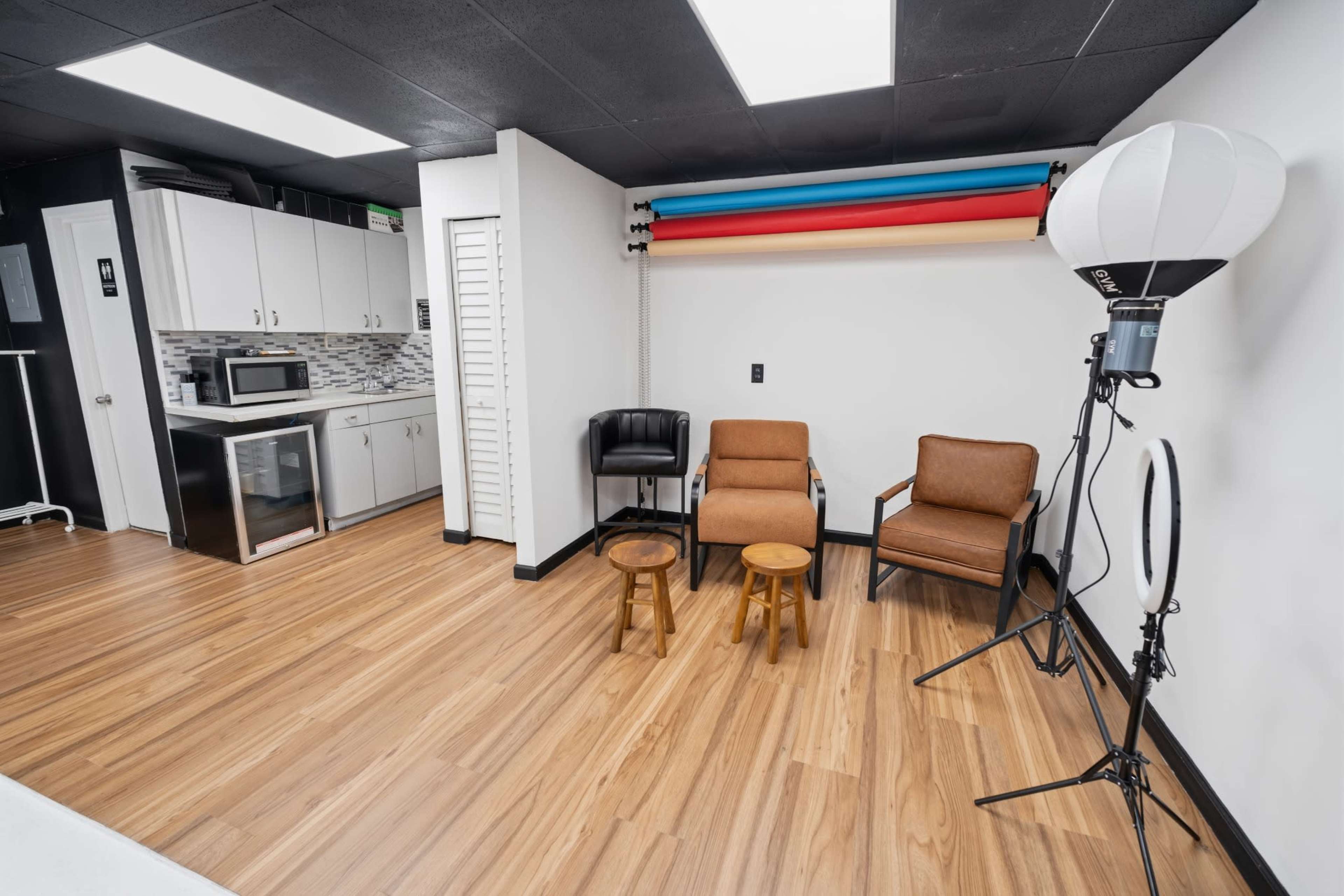 The image shows a well-lit studio space featuring a kitchenette area to the left, two brown chairs, and a photography backdrop setup on the right.