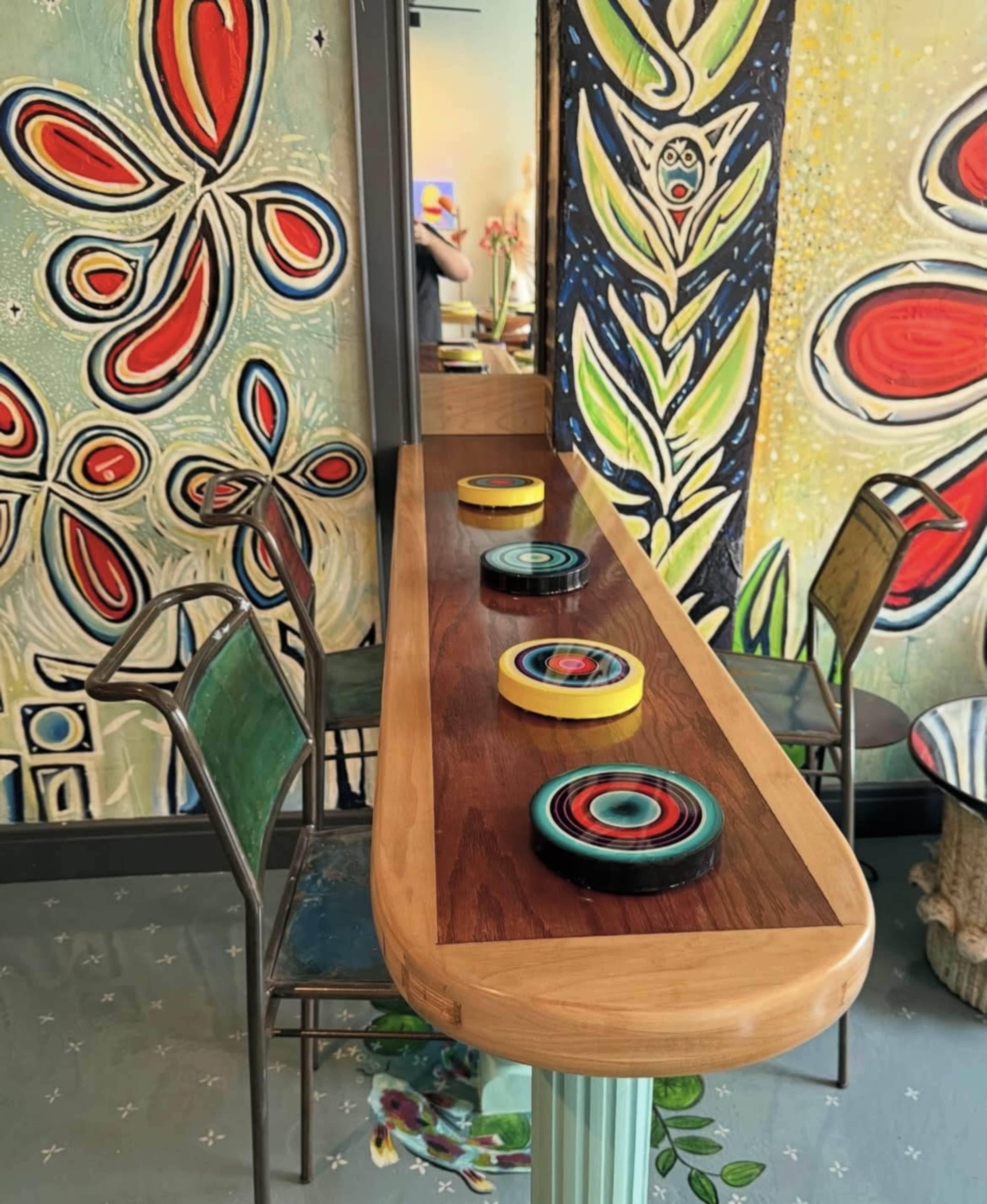 A long wooden table with colorful circular decorations sits against a vibrant wall adorned with abstract floral artwork, accompanied by two metal chairs.