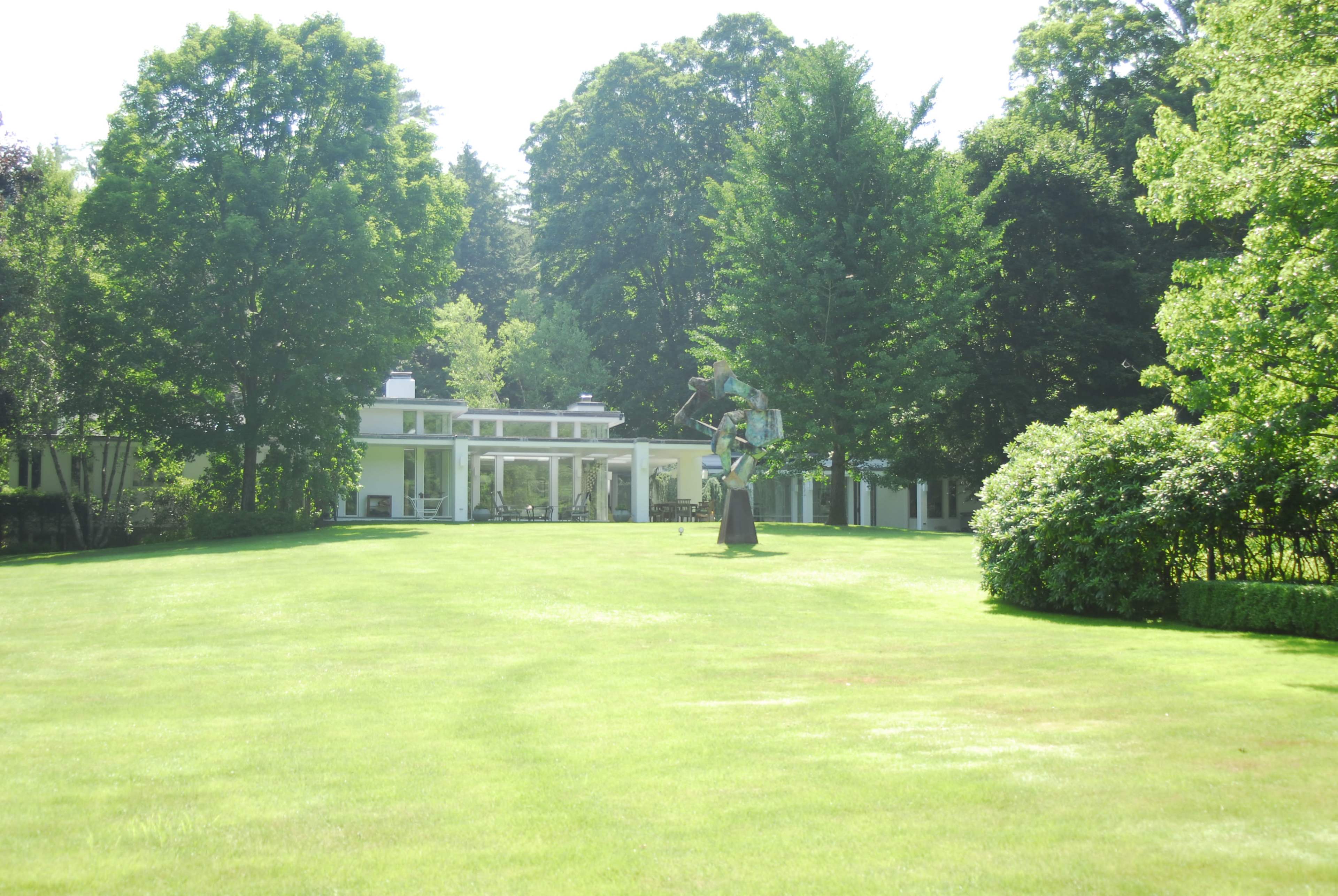 A large, manicured lawn leads to a modern house featuring large glass windows, with a sculpture situated on the grounds amidst trees.