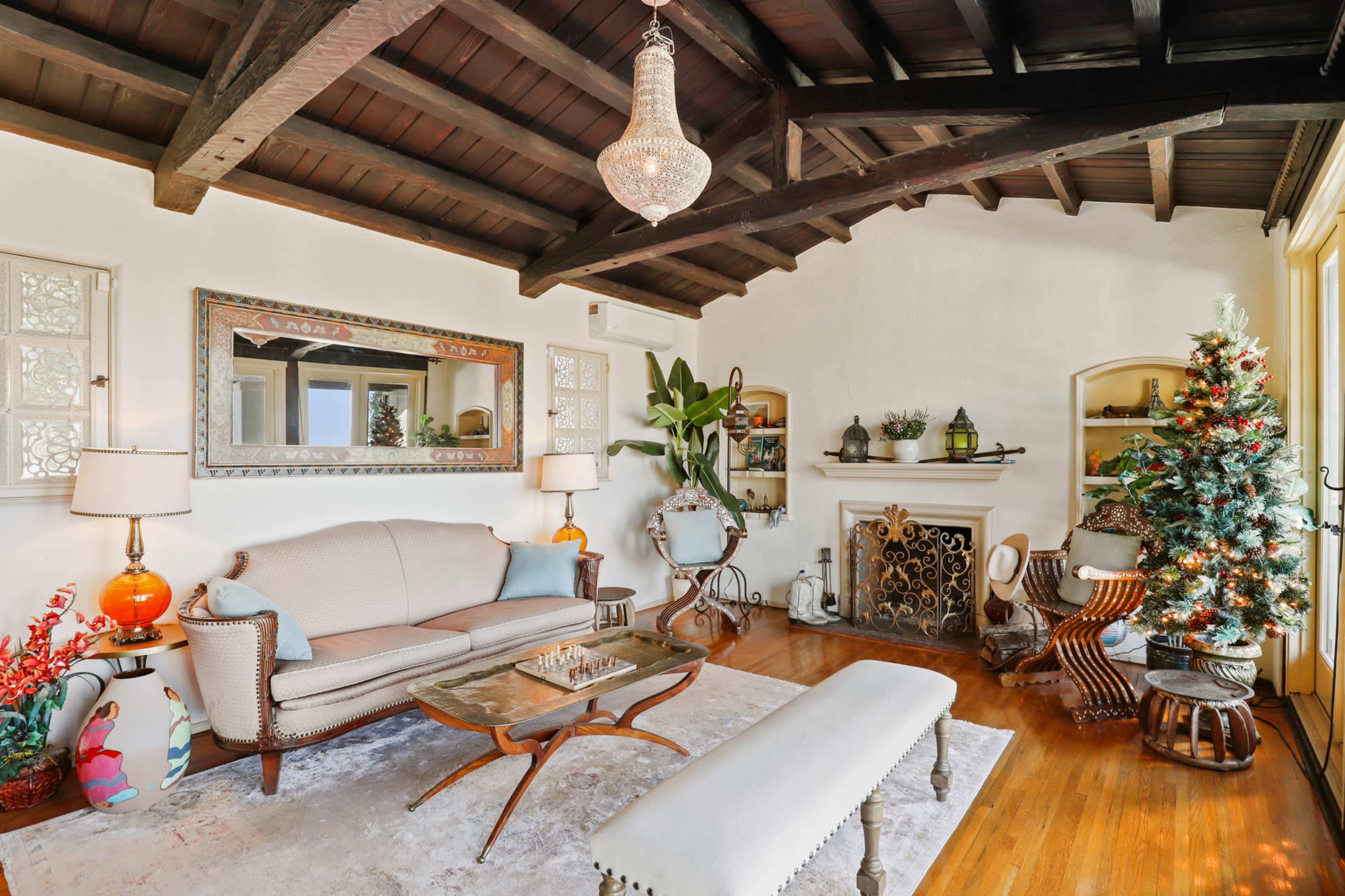 A cozy living room features a beige sofa, a glass coffee table, and a decorated Christmas tree beside a fireplace, complemented by wooden beams on the ceiling.