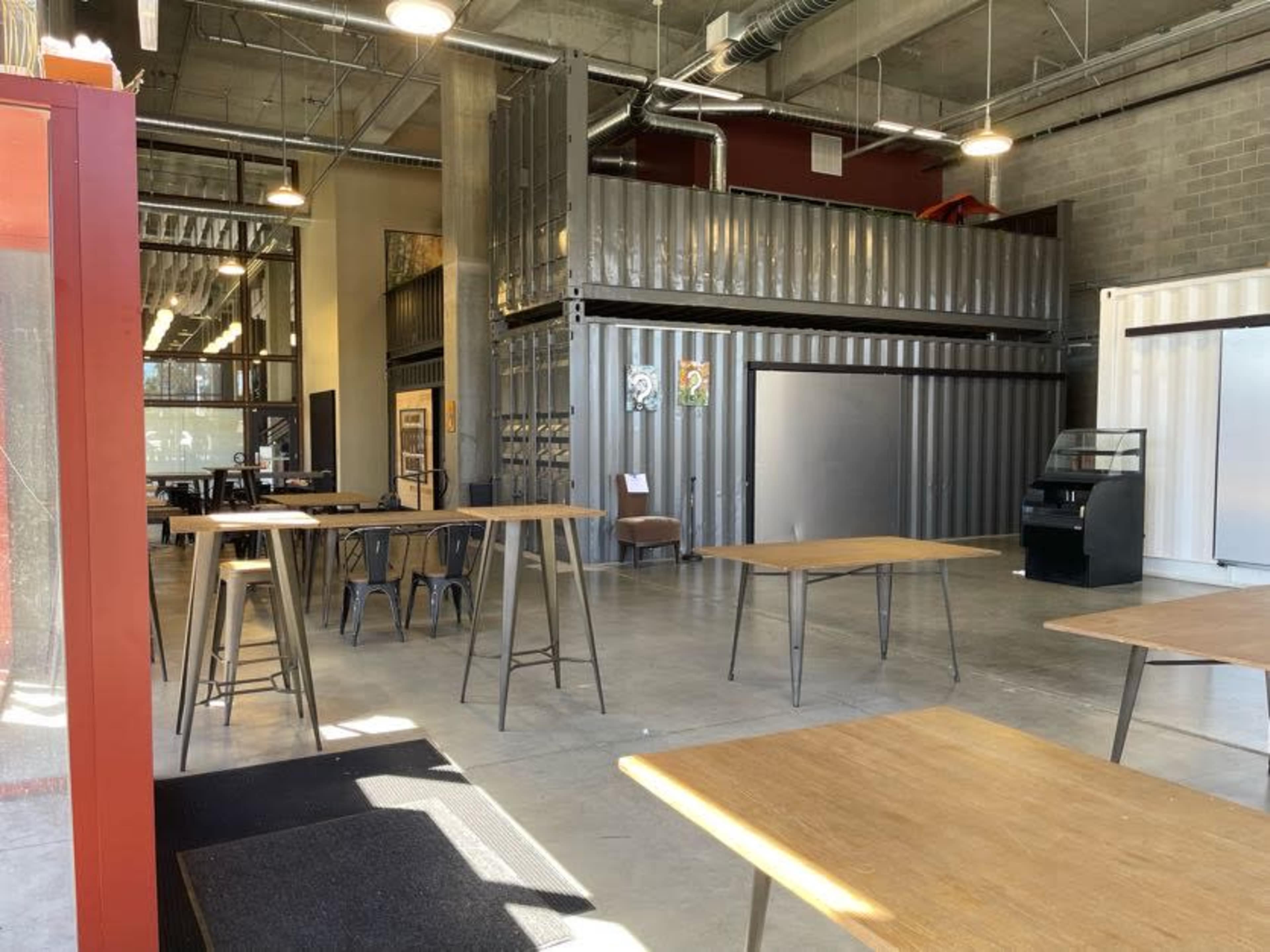 An industrial-style interior features metal tables and walls constructed from shipping containers in a spacious area with concrete flooring.