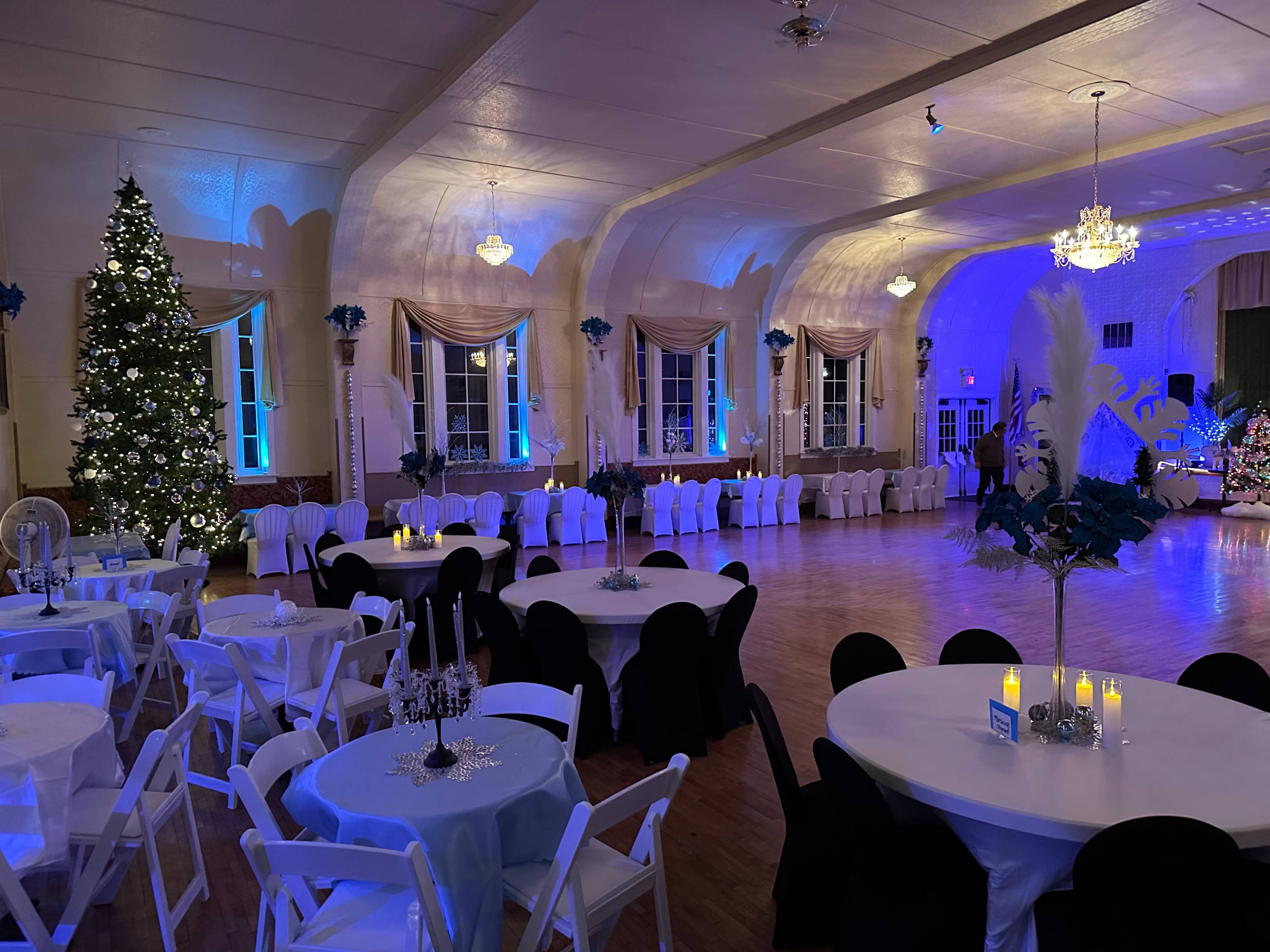A decorated reception hall with circular tables covered in white and black linens, a large Christmas tree in the corner, and soft blue lighting illuminating the space.
