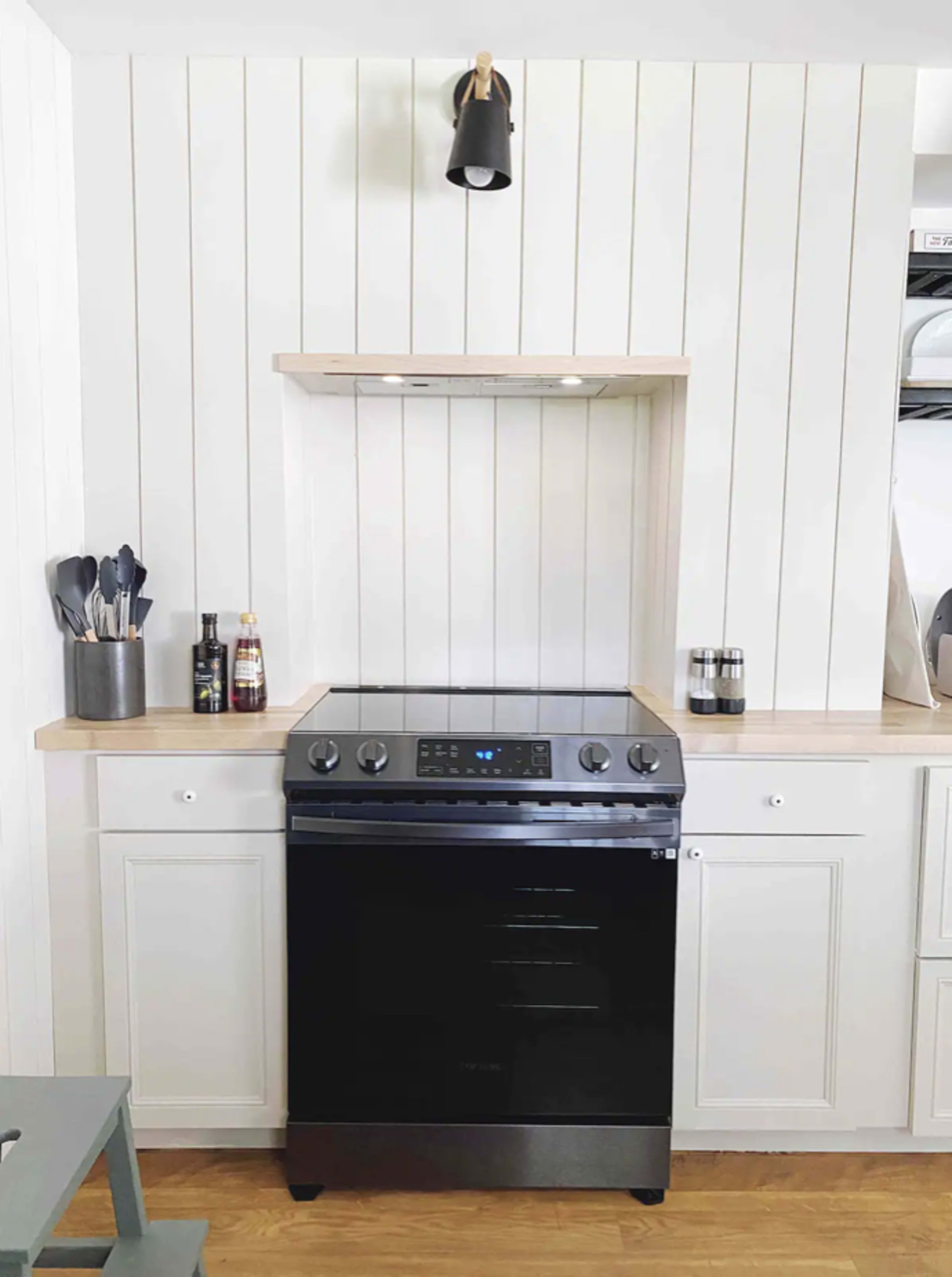 A modern kitchen setup featuring a black stove beneath a wooden shelf, with white shiplap walls and light wood countertops.