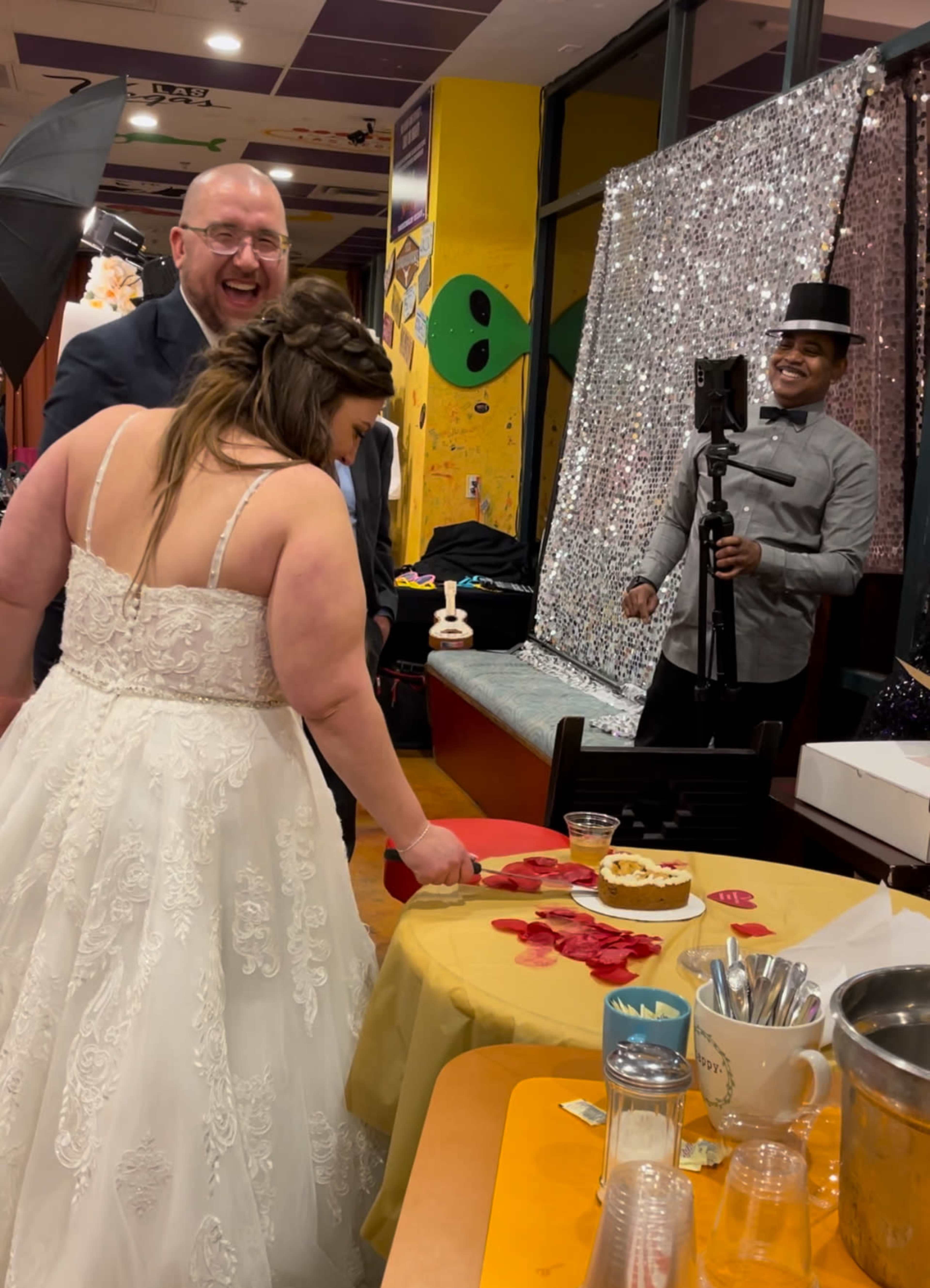 A bride in a white gown laughs as she prepares to cut a cake at a decorated table, while a man in a suit and another in a top hat look on.