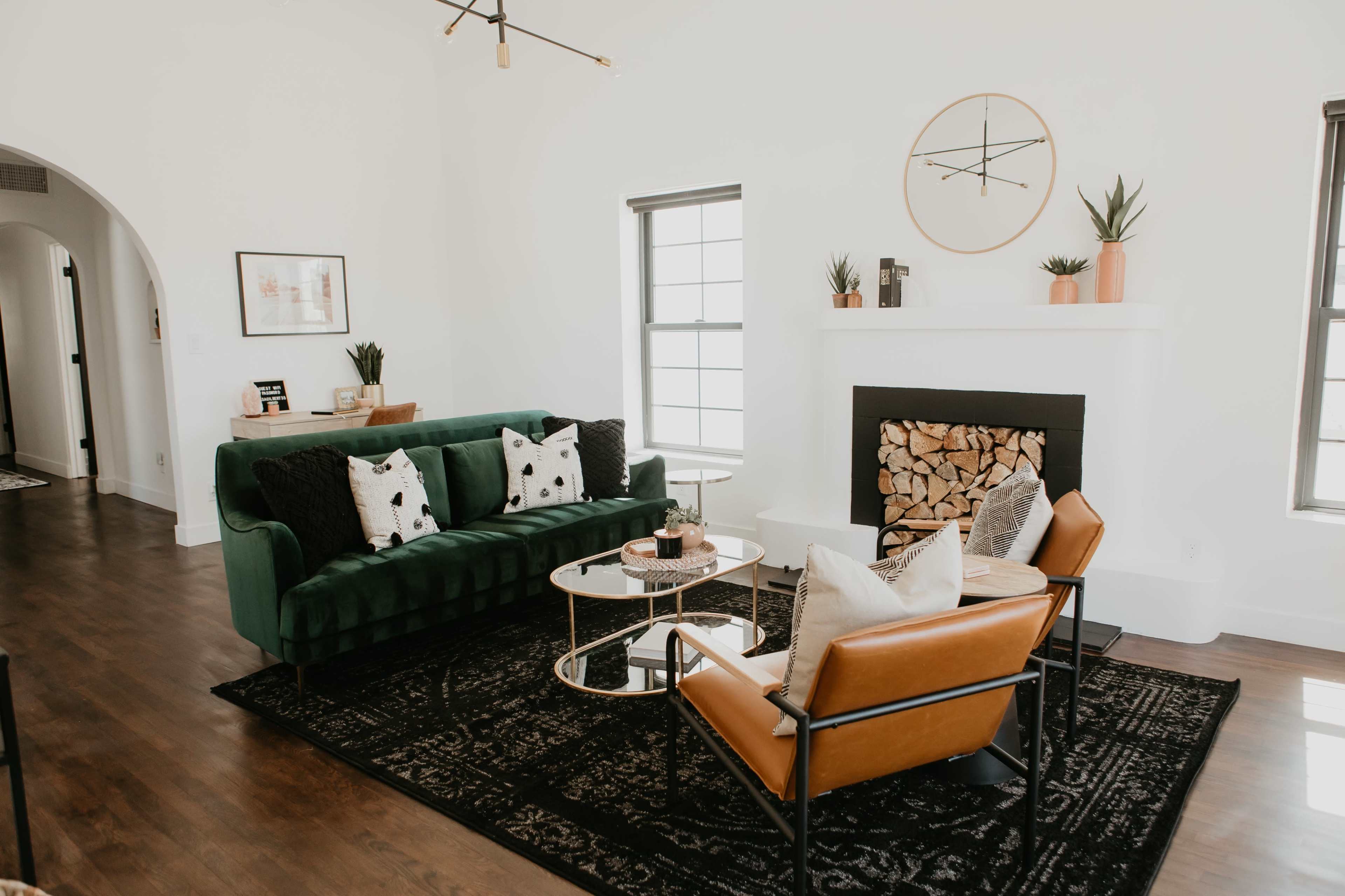 The living room features a green velvet sofa, a brown leather chair, a coffee table, and a fireplace adorned with stacked wood, all against a backdrop of white walls.