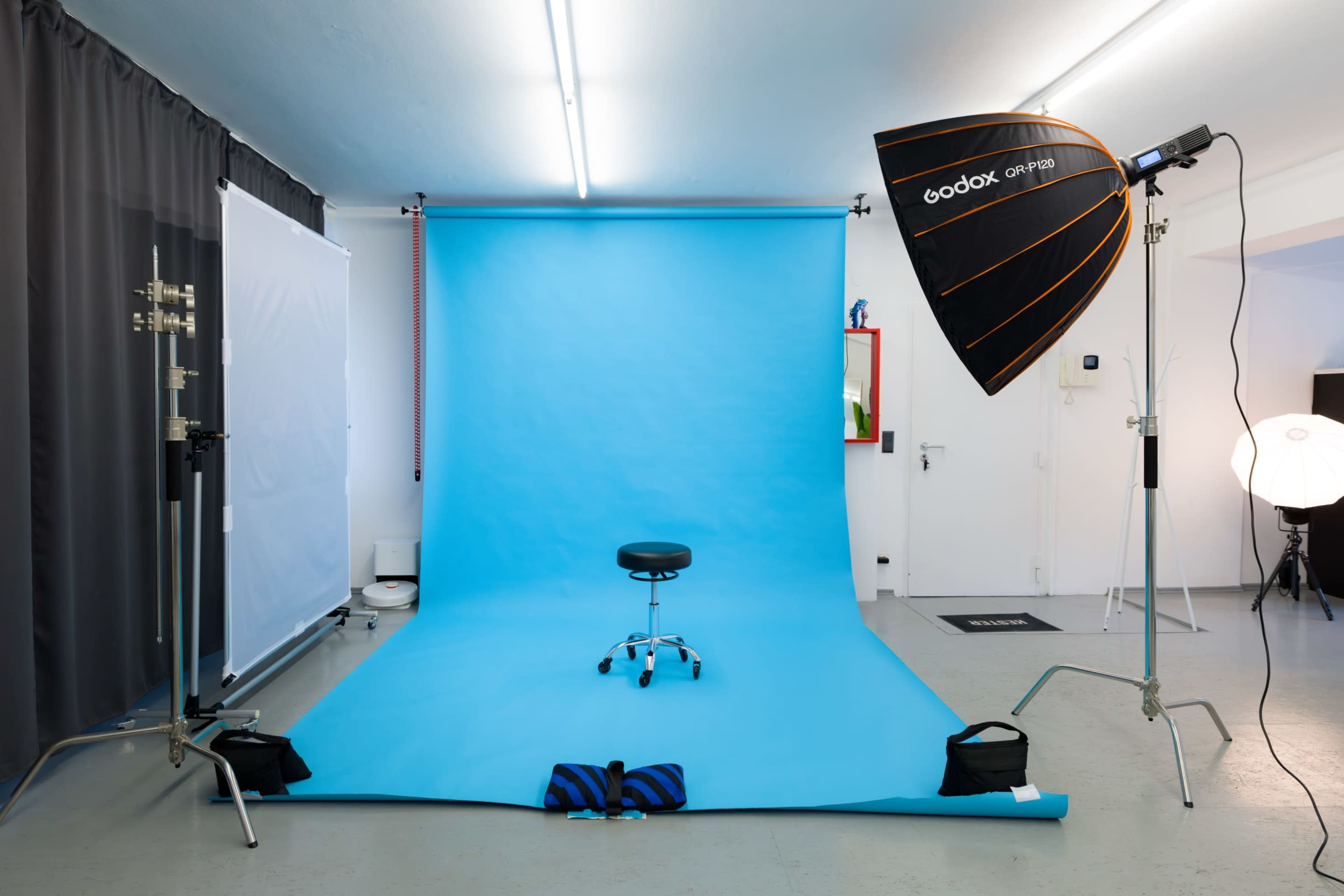 The image depicts a photography studio with a blue backdrop, a stool in the center, and studio lighting equipment set up around the area.
