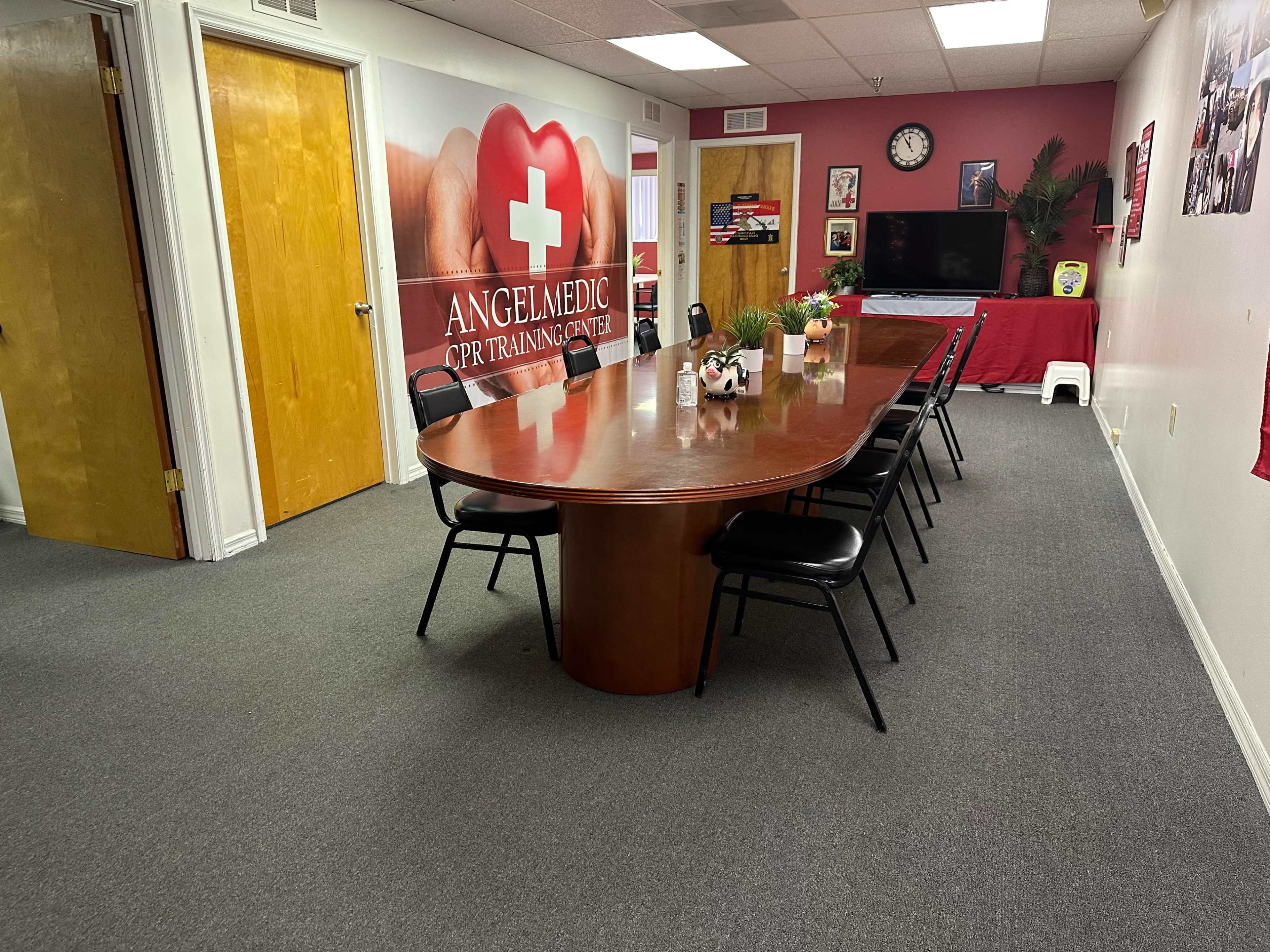 A training room features a long wooden conference table surrounded by black chairs, with a wall displaying a large red cross and medical images.