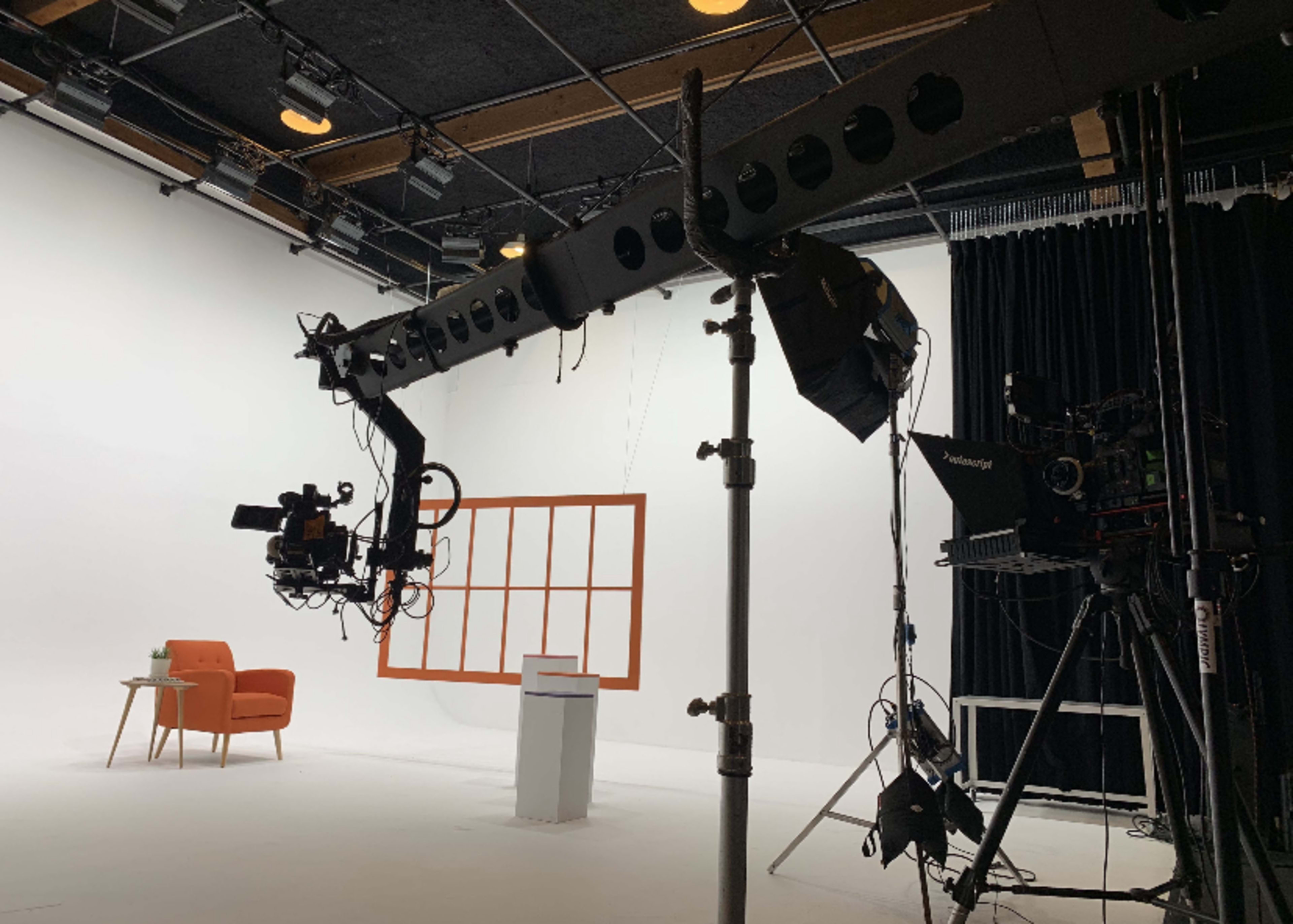 The image shows a film set with a camera crane, lighting equipment, and an orange armchair in front of a white backdrop.