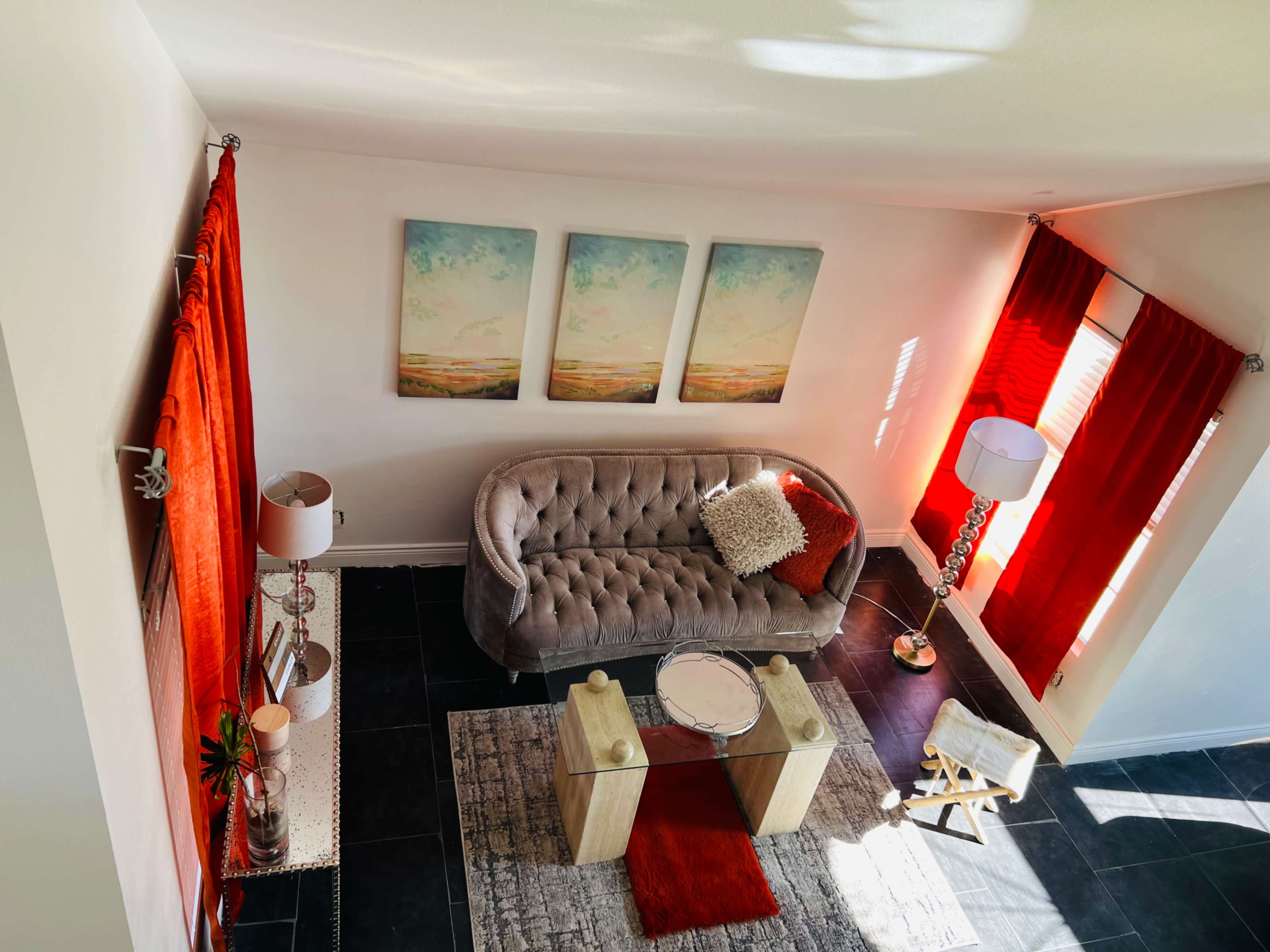 A stylish living room with a gray tufted sofa, a round glass coffee table, and vibrant red curtains framing two windows.
