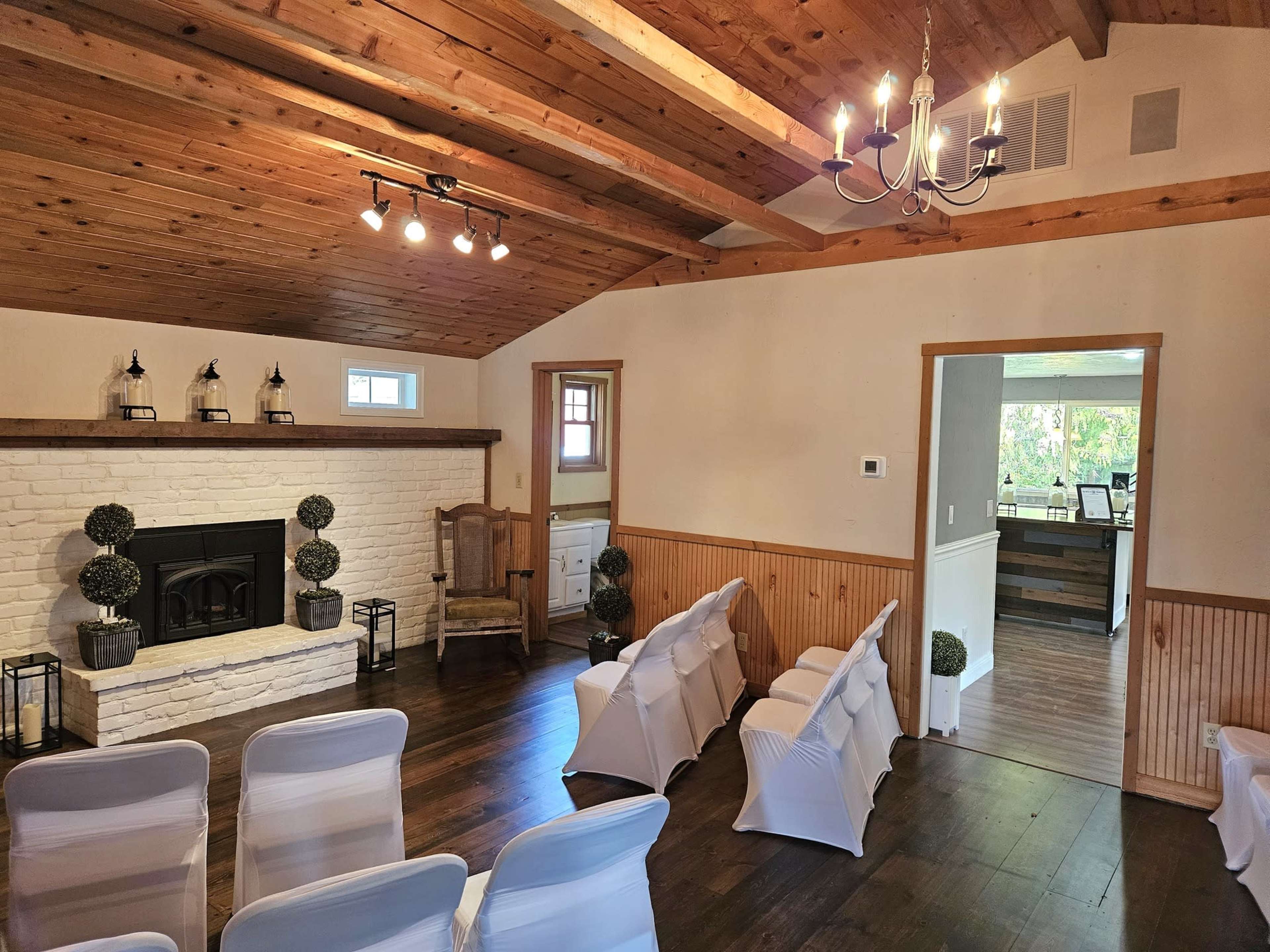An indoor ceremony space featuring white chairs arranged in front of a brick fireplace, with wooden beams on the ceiling and a table visible in the adjacent room.