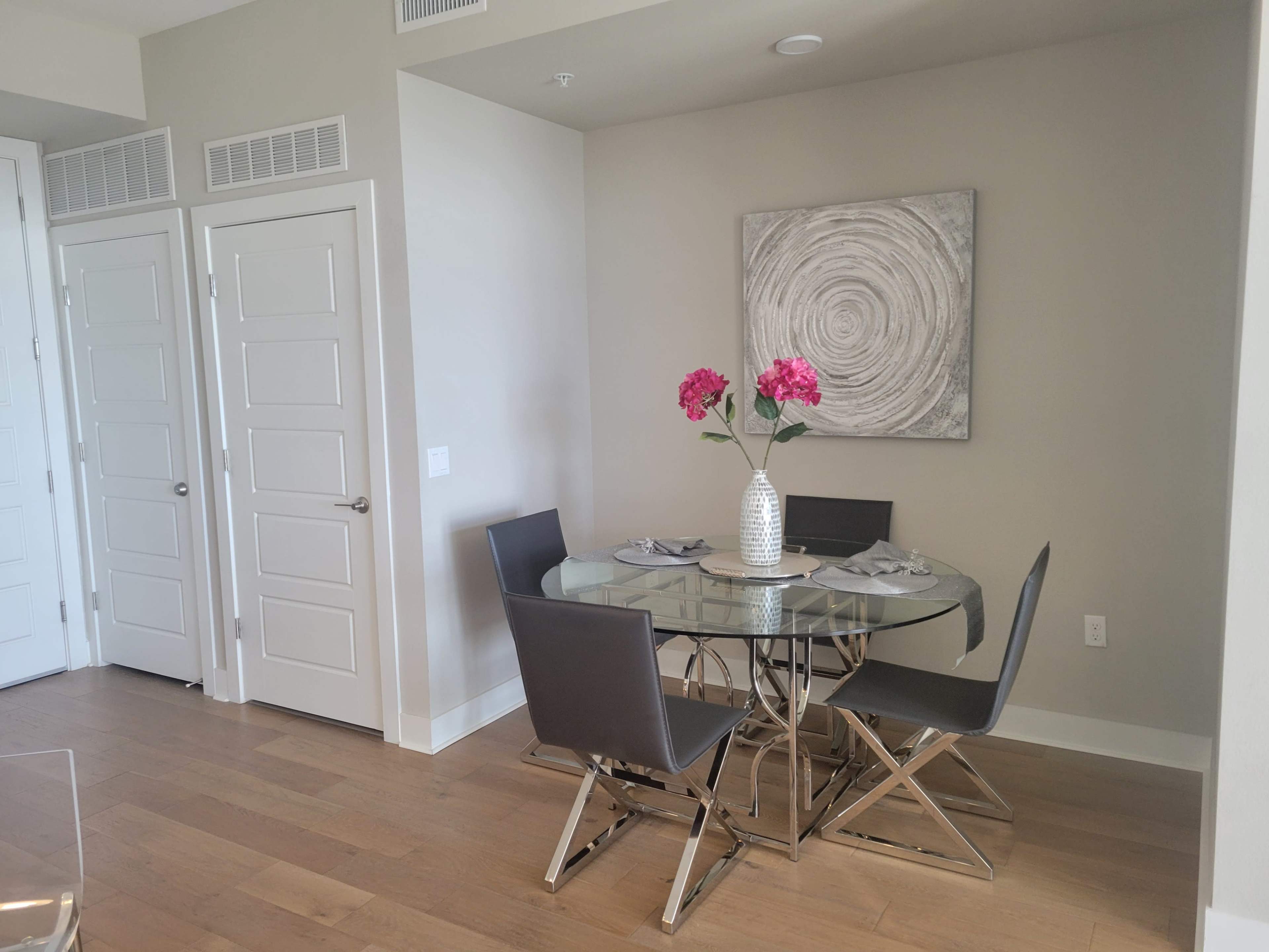 A glass dining table surrounded by four black chairs is set with flowers in a vase against a neutral wall with a circular artwork.