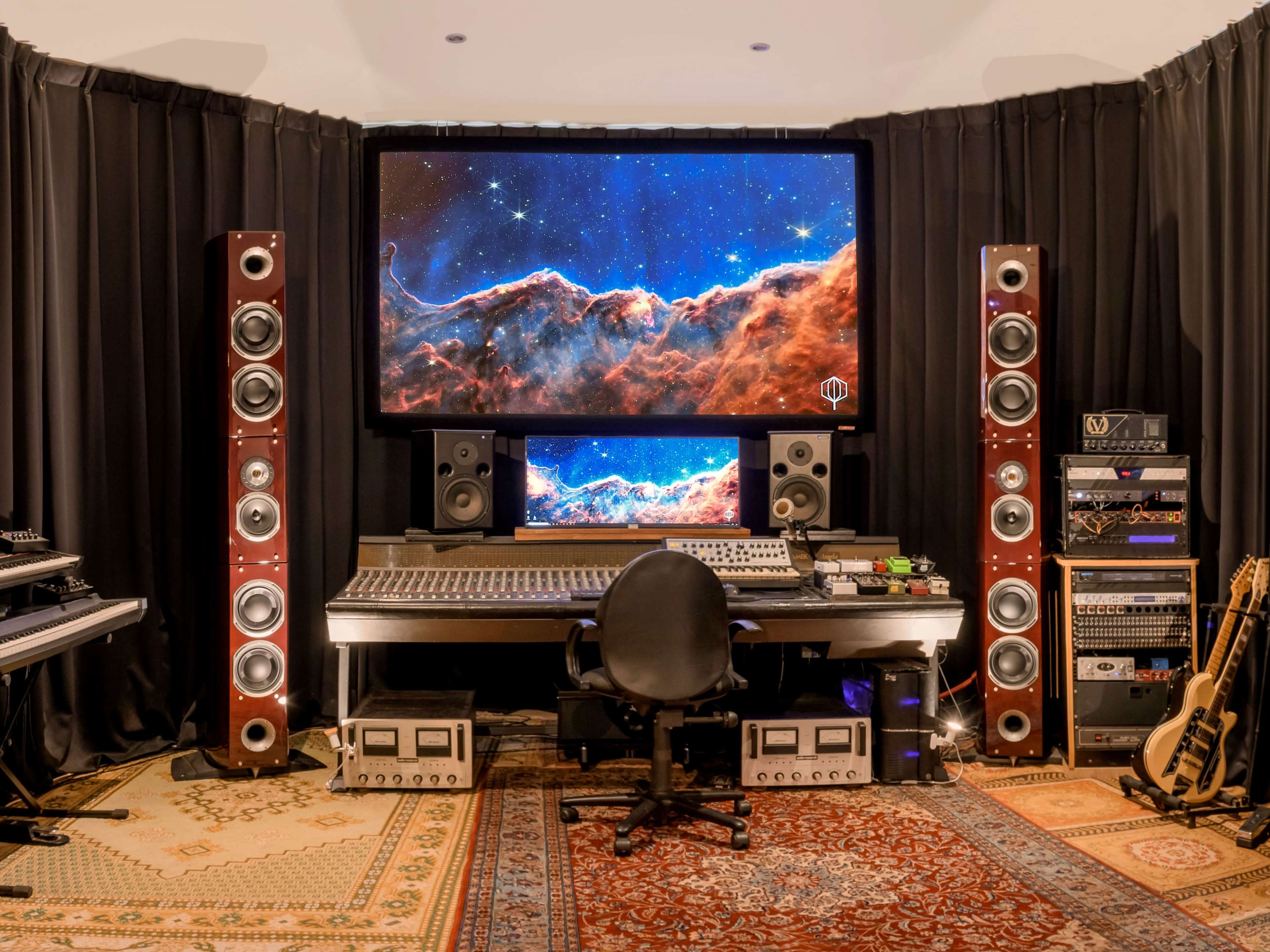 A modern music production studio features a large central workstation with two screens displaying a cosmic scene, surrounded by tall speakers and various music equipment.