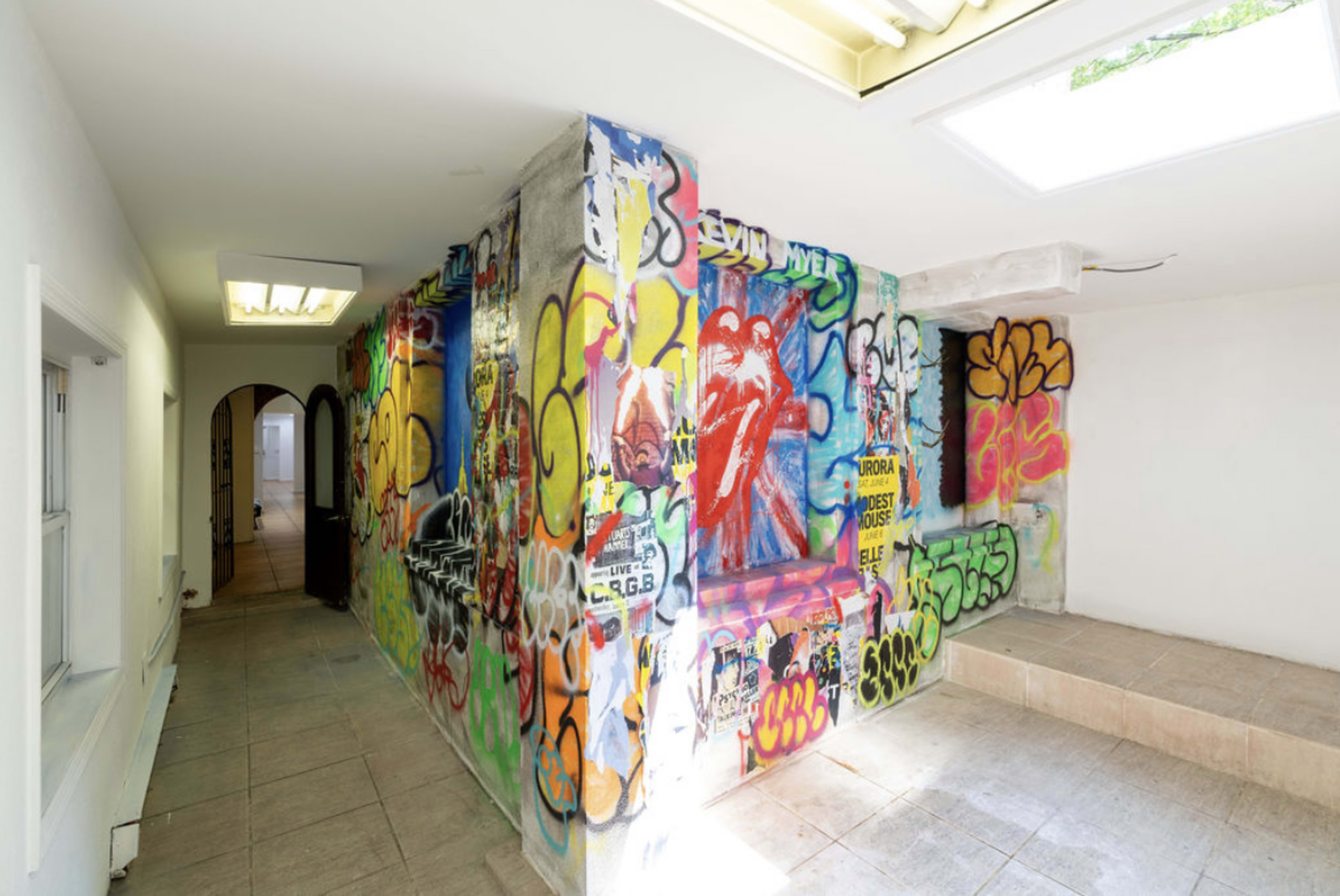 A hallway with walls covered in vibrant graffiti and street art, contrasting with the plain tiled floor and ceiling.