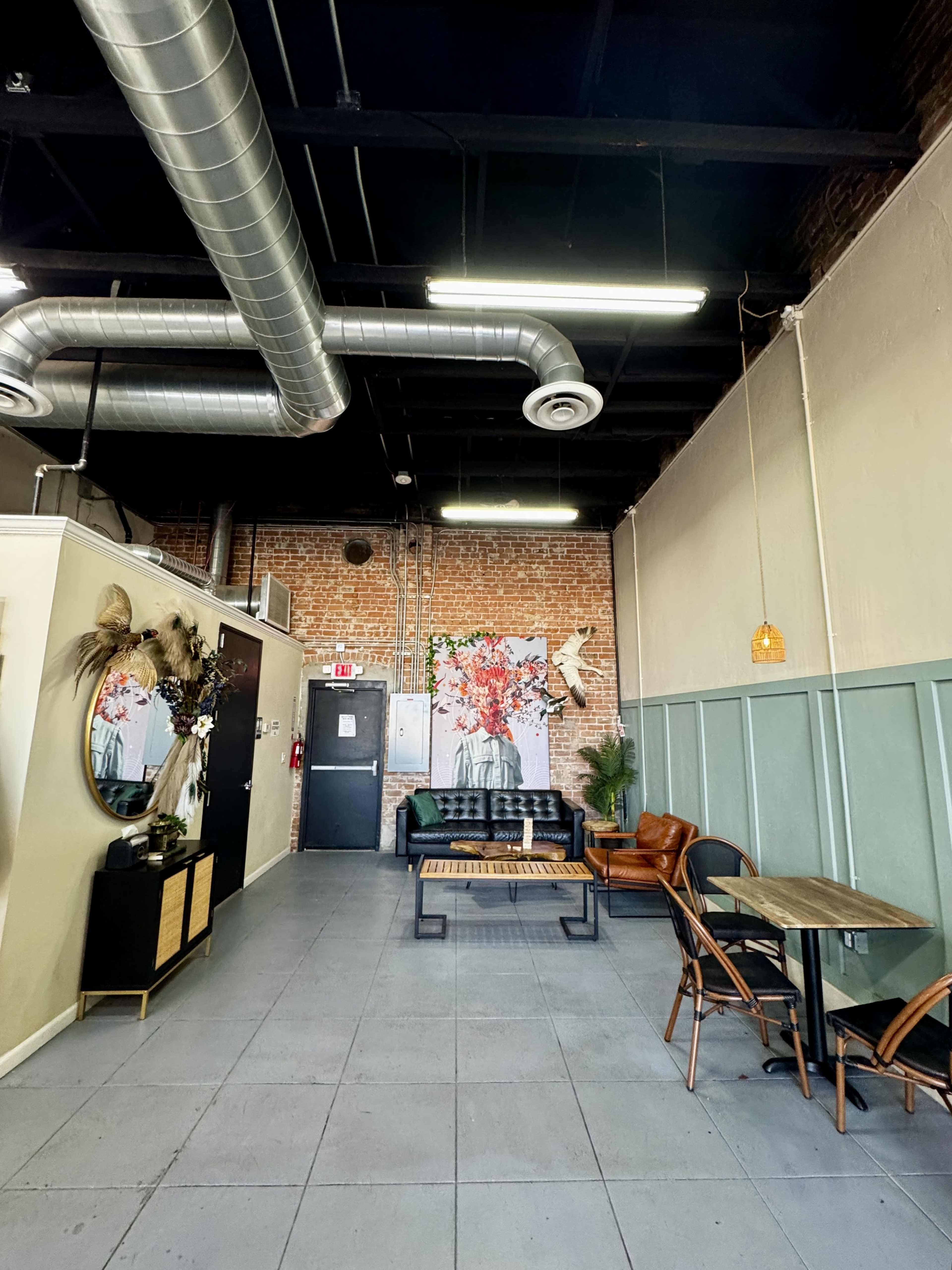 The image shows a modern interior space with exposed brick walls, a floral mural, and a seating area that includes a black sofa and wooden tables.