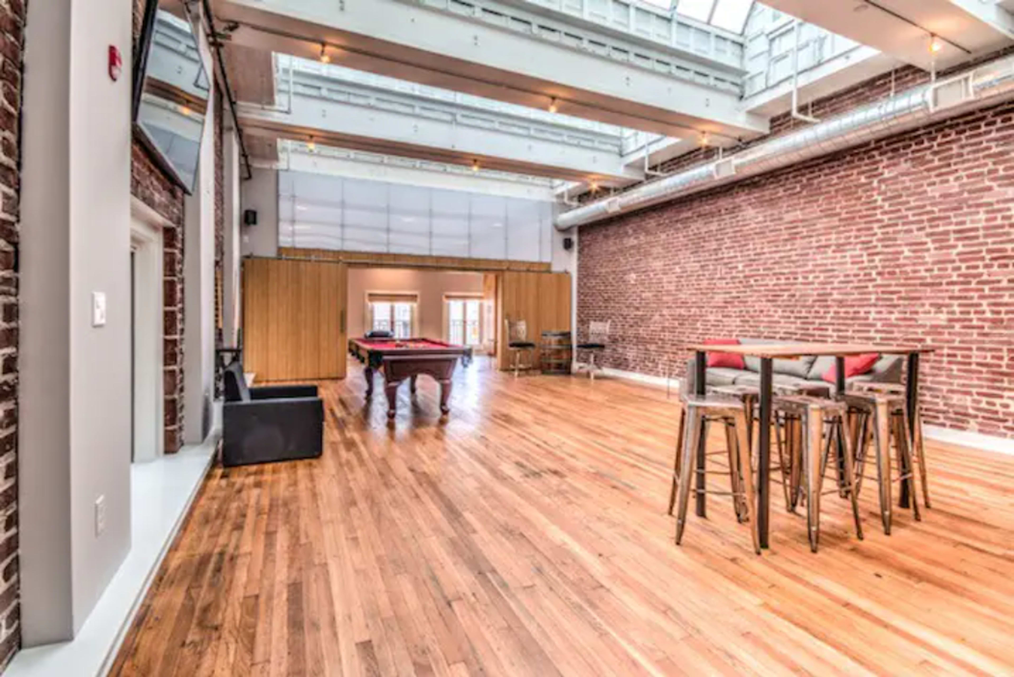 A spacious, modern living area featuring exposed brick walls, wooden flooring, and a pool table in the background.