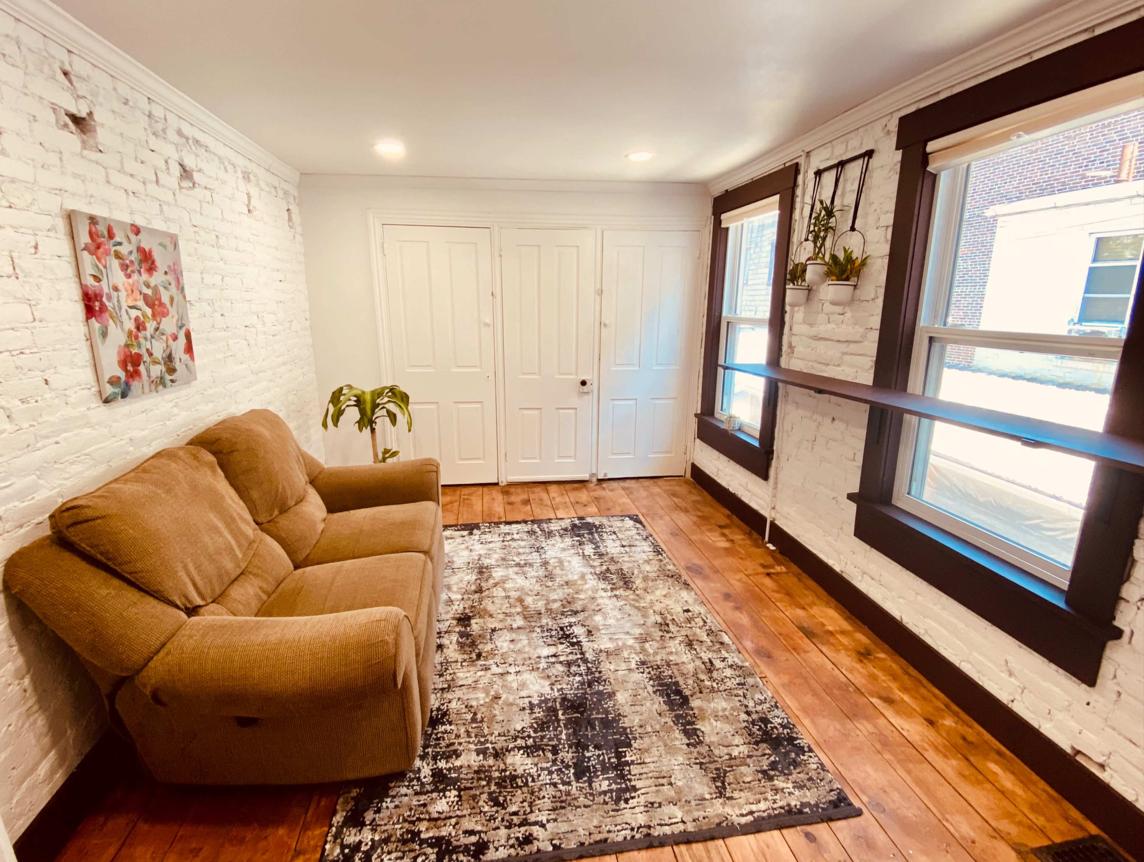A cozy living room with a brown couch, a patterned rug, and brick-walled accents, featuring two windows and a pair of closed white doors.