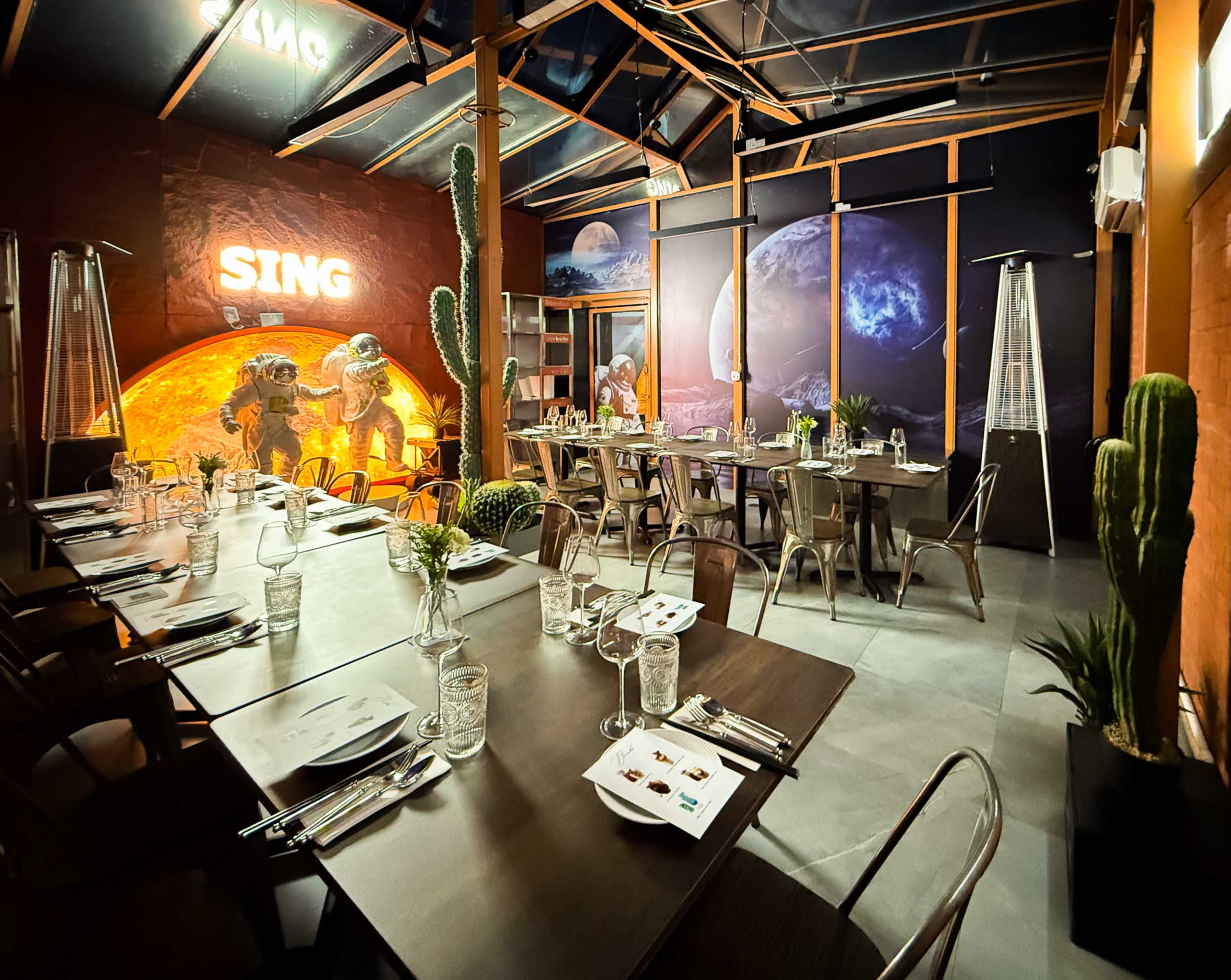 The image shows an indoor restaurant setting with long tables, metal chairs, and a large wall mural depicting a space scene with planets and astronauts.
