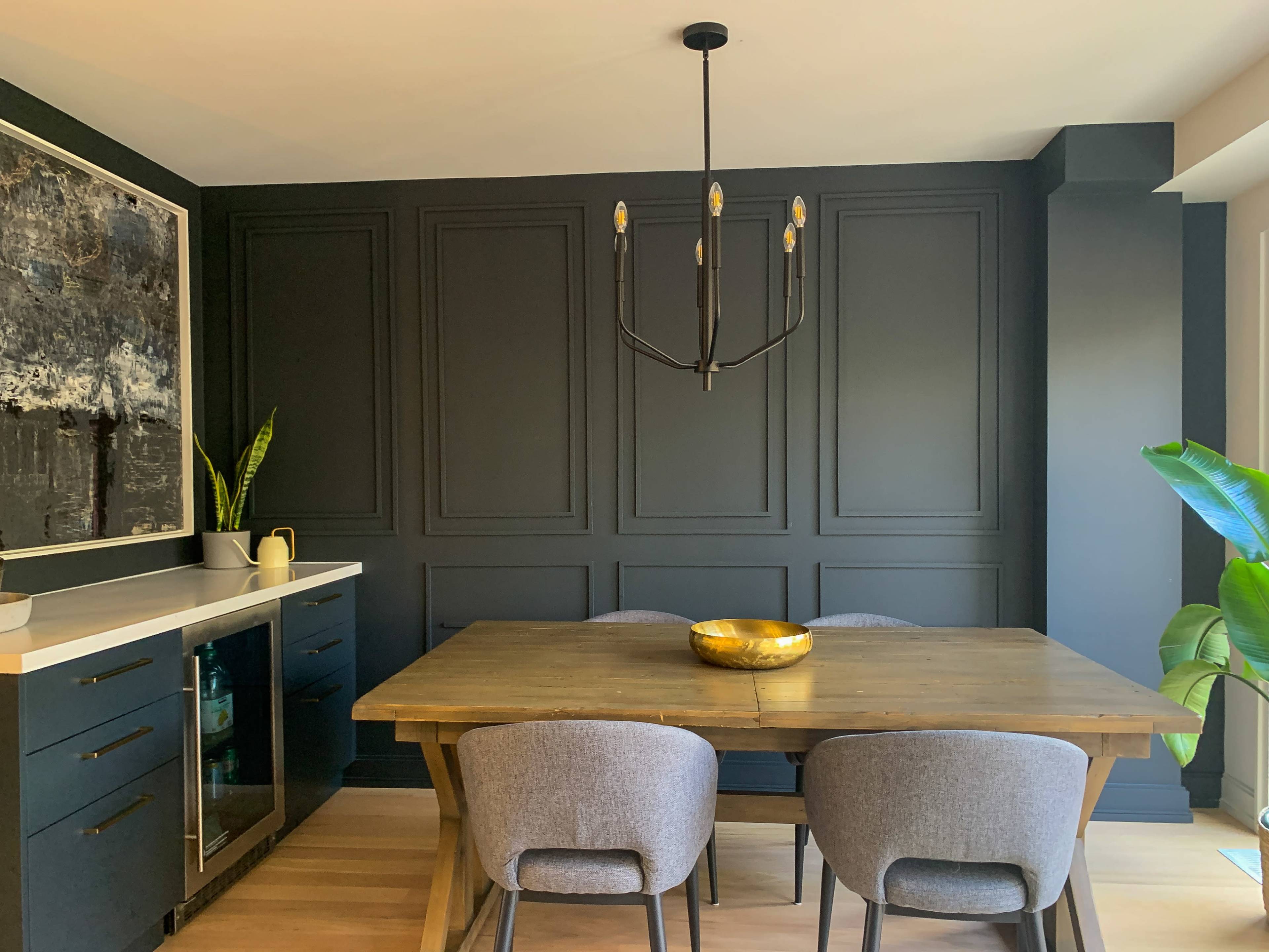 A dining area features a wooden table surrounded by gray chairs, a pendant light hanging above, and black paneled walls adorned with a large artwork.