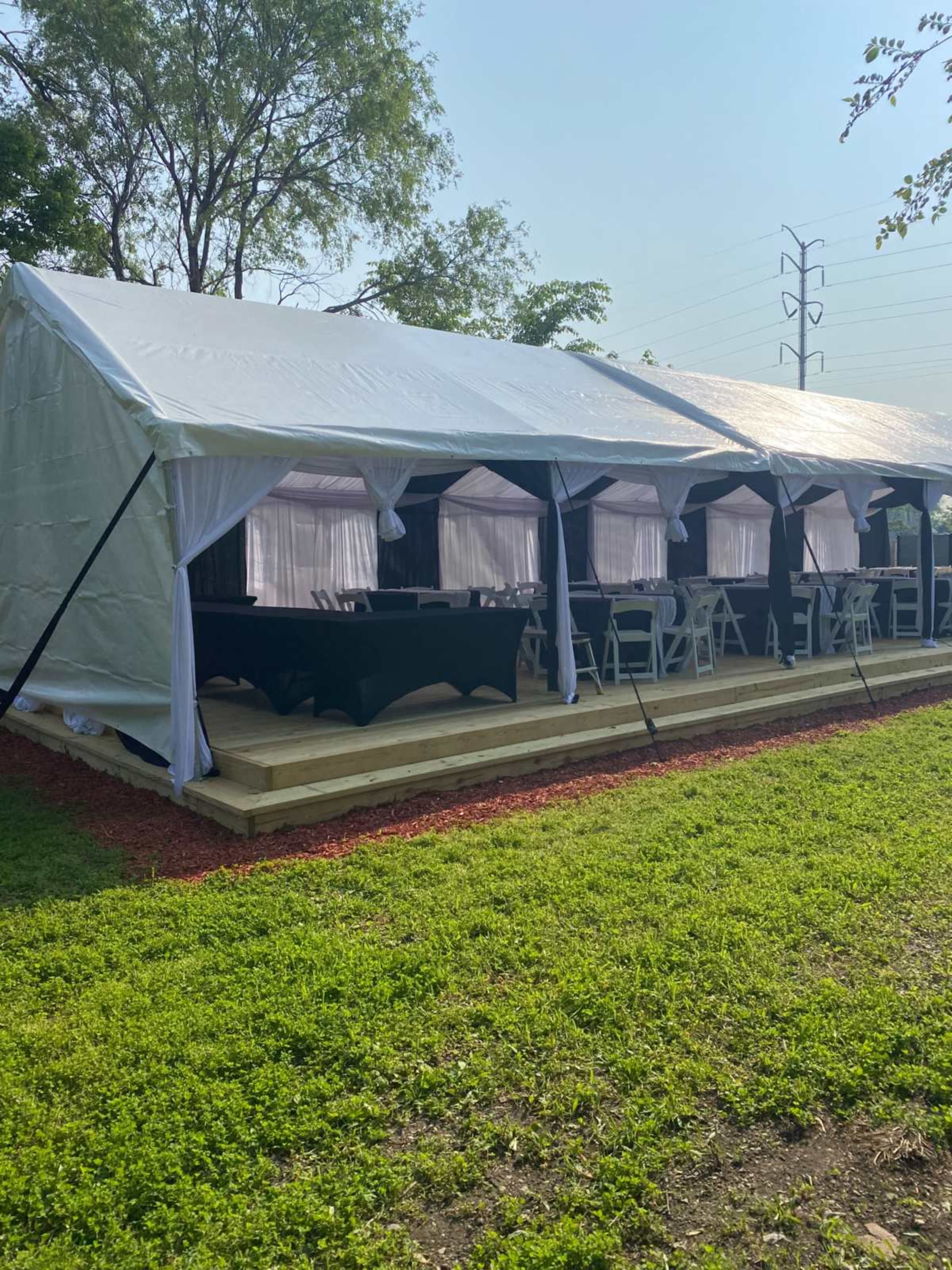 A large white tent with open sides sits on a grassy area, featuring multiple tables covered with black tablecloths and surrounded by chairs.