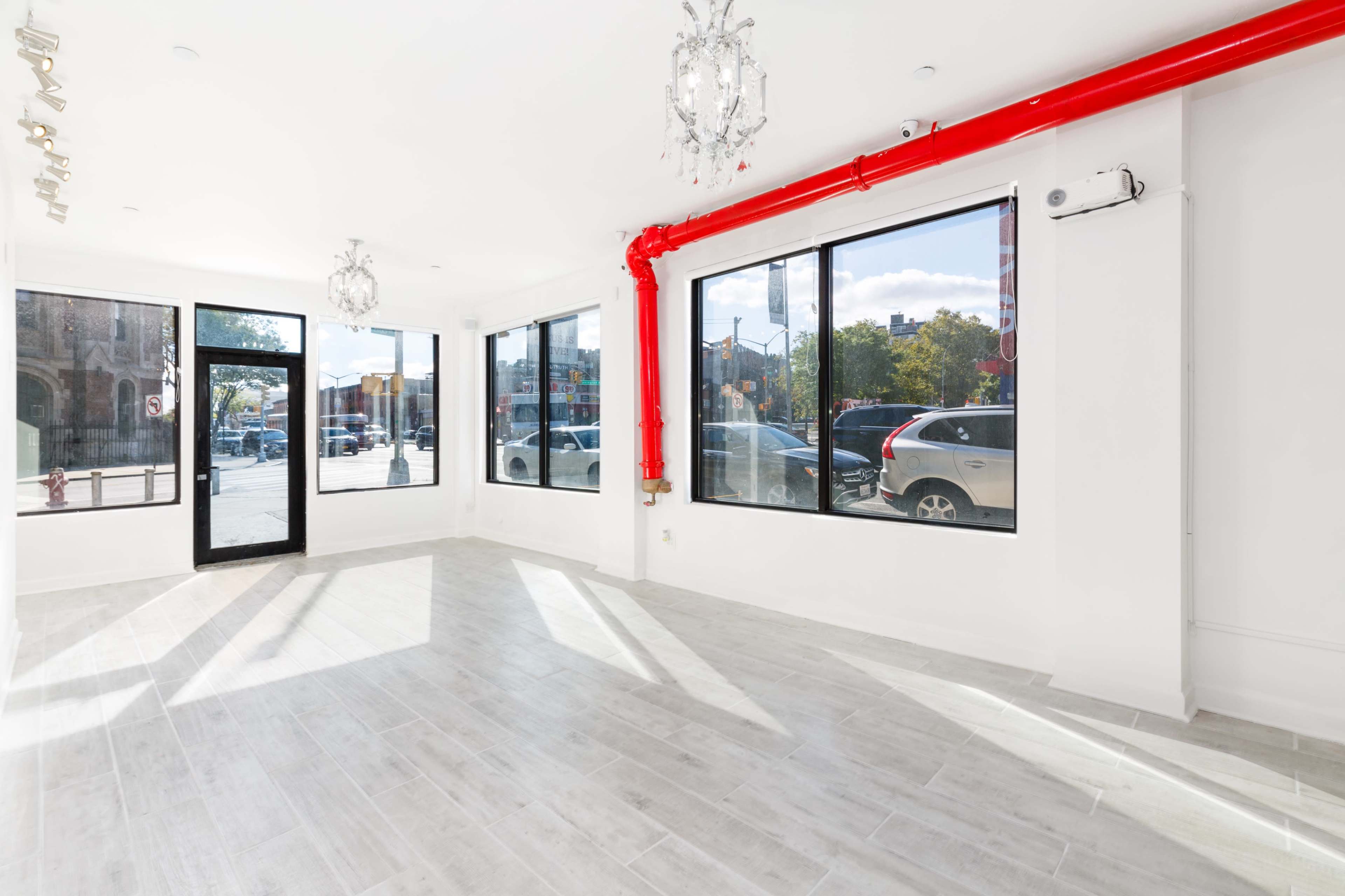 An empty retail space with large windows, white walls, and grey tile flooring, featuring red piping along the ceiling.