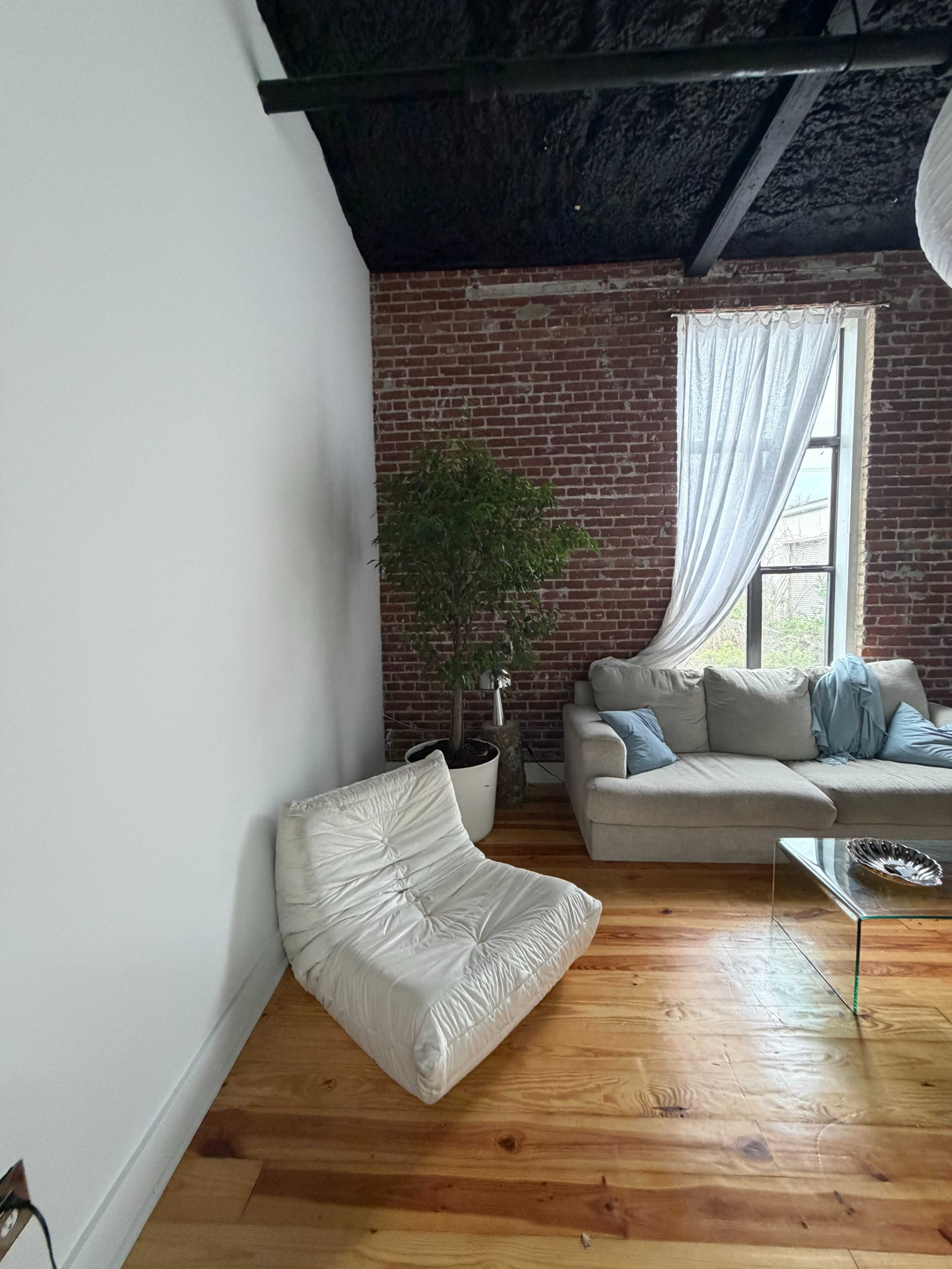 The image shows a modern living room with a brick wall, a large window covered by sheer curtains, a light-colored sofa, a small black bean bag chair, and a wooden floor.