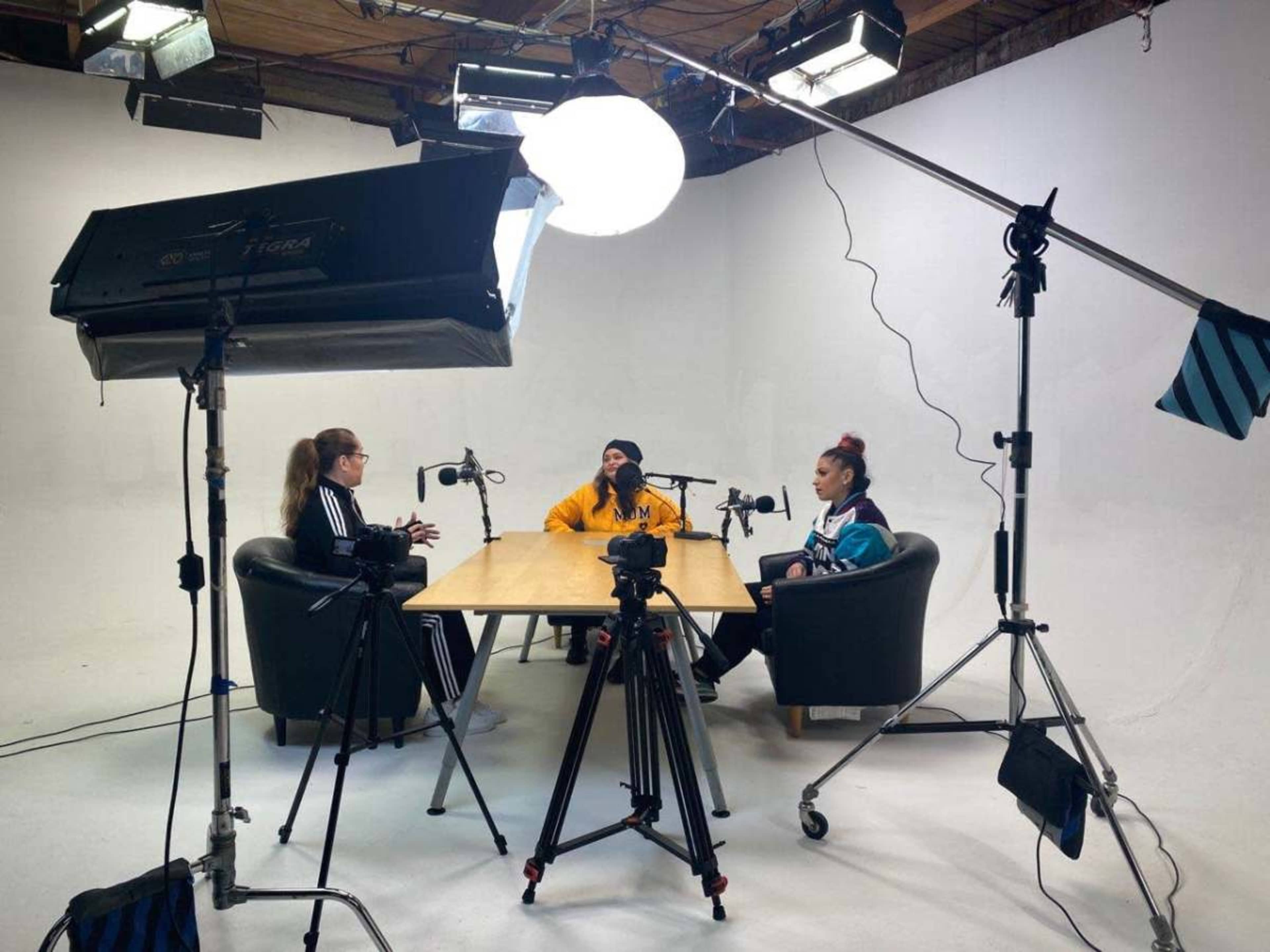 Three people sit at a table in a studio setting, surrounded by microphones and lighting equipment, while cameras are set up to capture the scene.