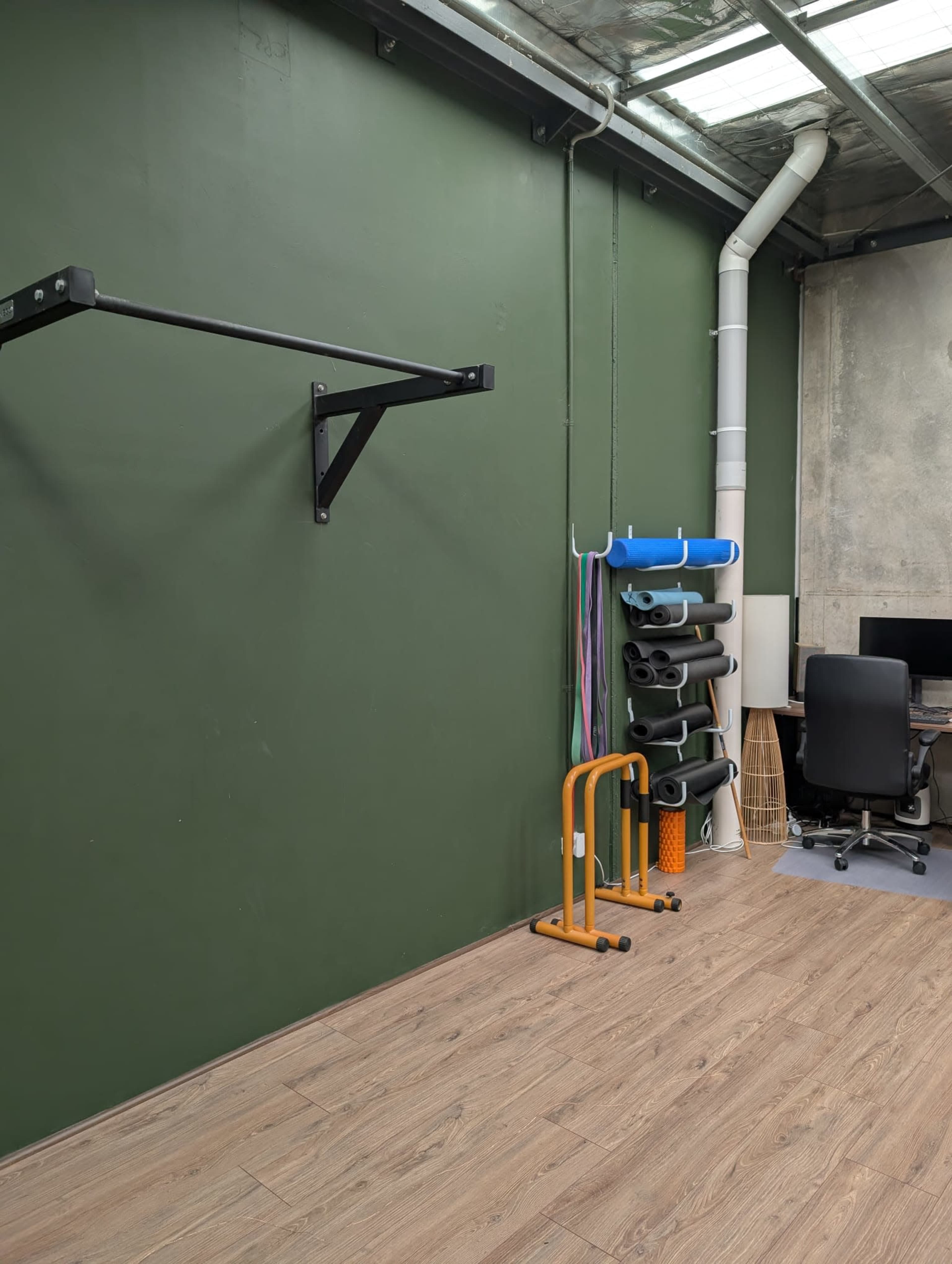 The image shows a minimalist gym space featuring a green wall with a pull-up bar, exercise mats stacked vertically, and a desk with an office chair in the background.