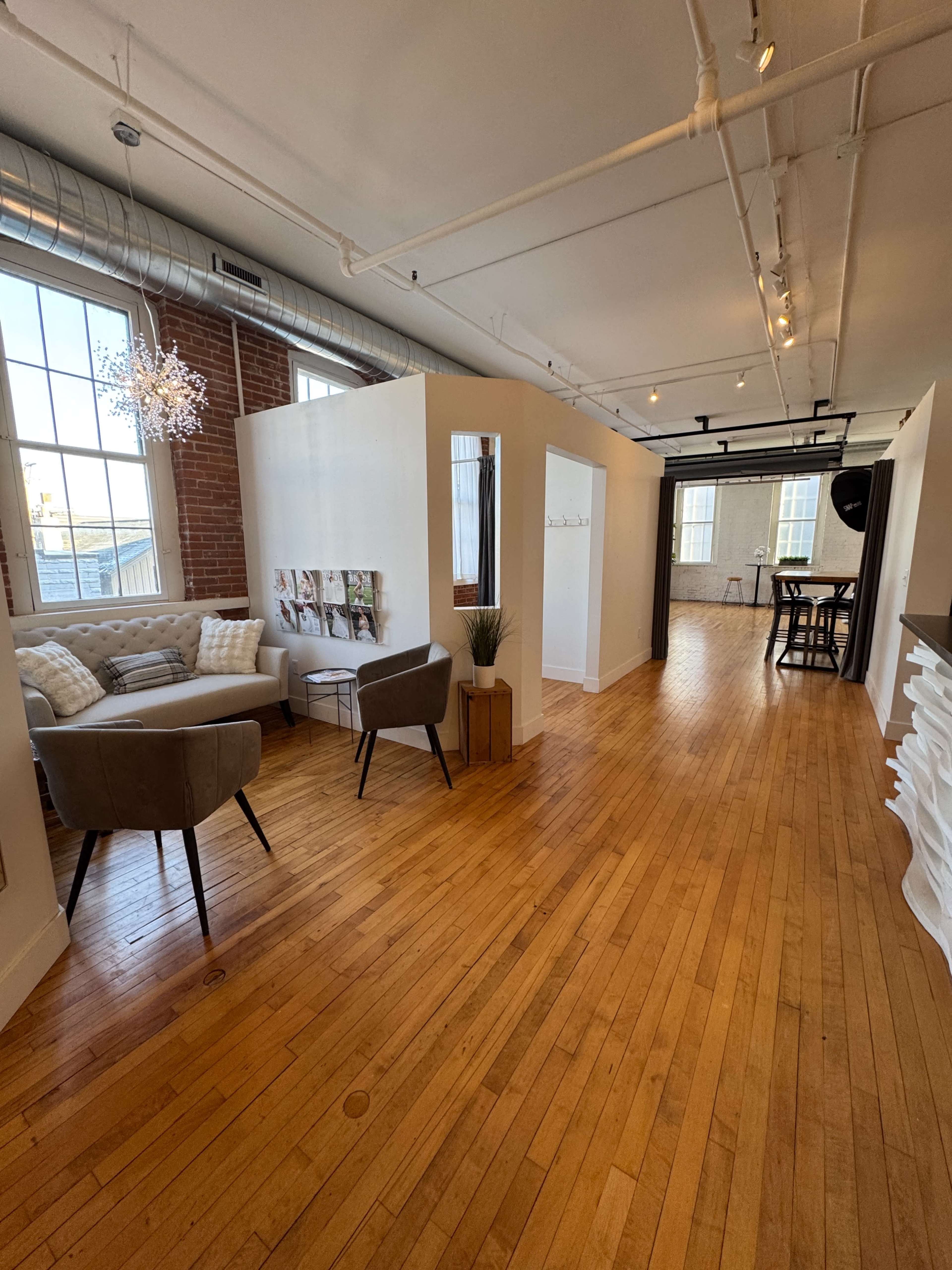 The image shows a spacious, modern interior with wooden floors, featuring a seating area with two armchairs, a small decorative table, and partitions creating distinct spaces.