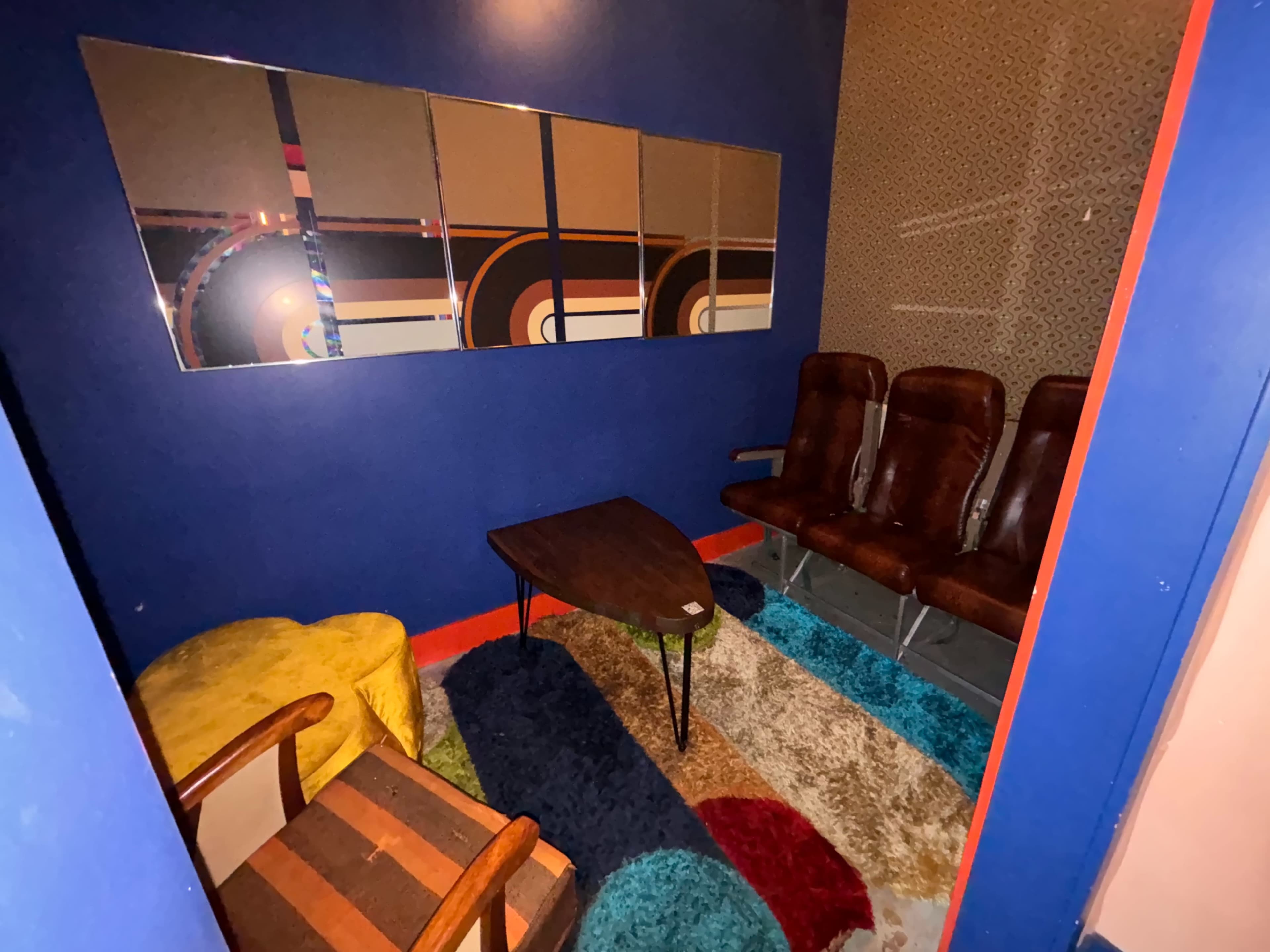 The image shows a cozy, small room with a blue wall featuring a coffee table, two armchairs, and several brown seating chairs arranged around a colorful rug.