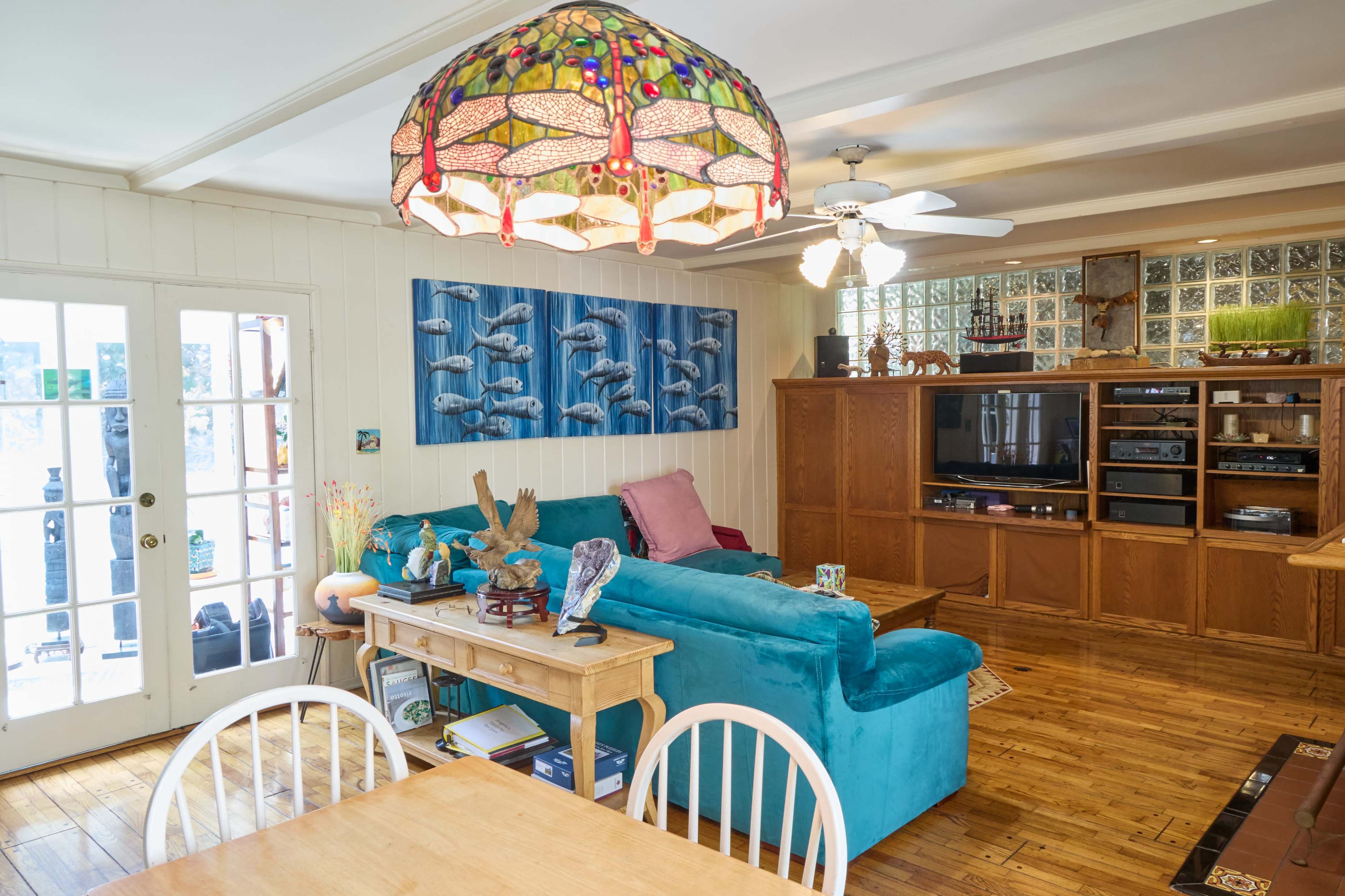 The image shows a cozy living room featuring a blue sofa, wooden furniture, a ceiling fan, and colorful decor, including a stained glass lamp.