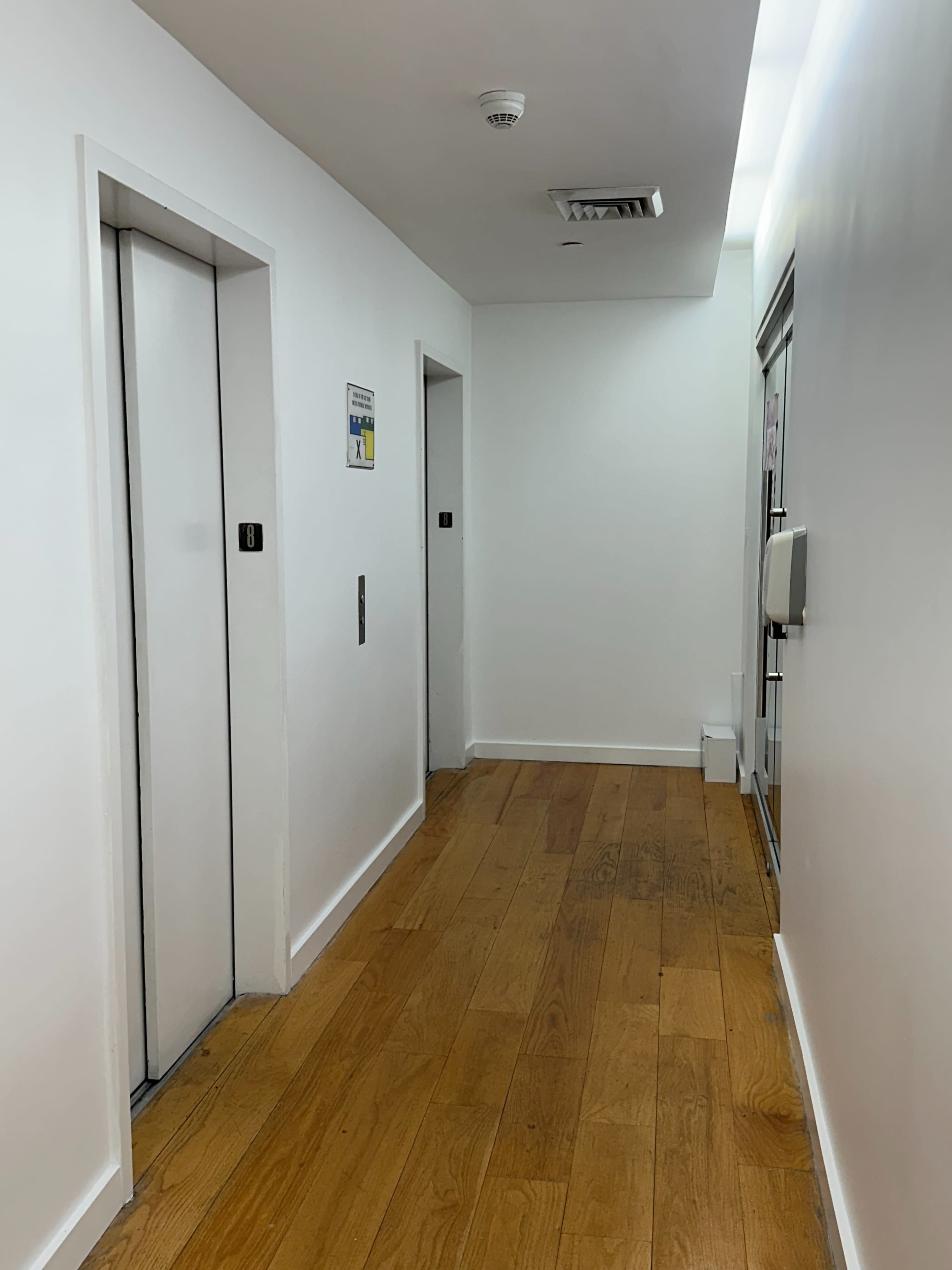 The image shows a long hallway with two elevator doors on the left and a glass door at the end, featuring wooden flooring and white walls.