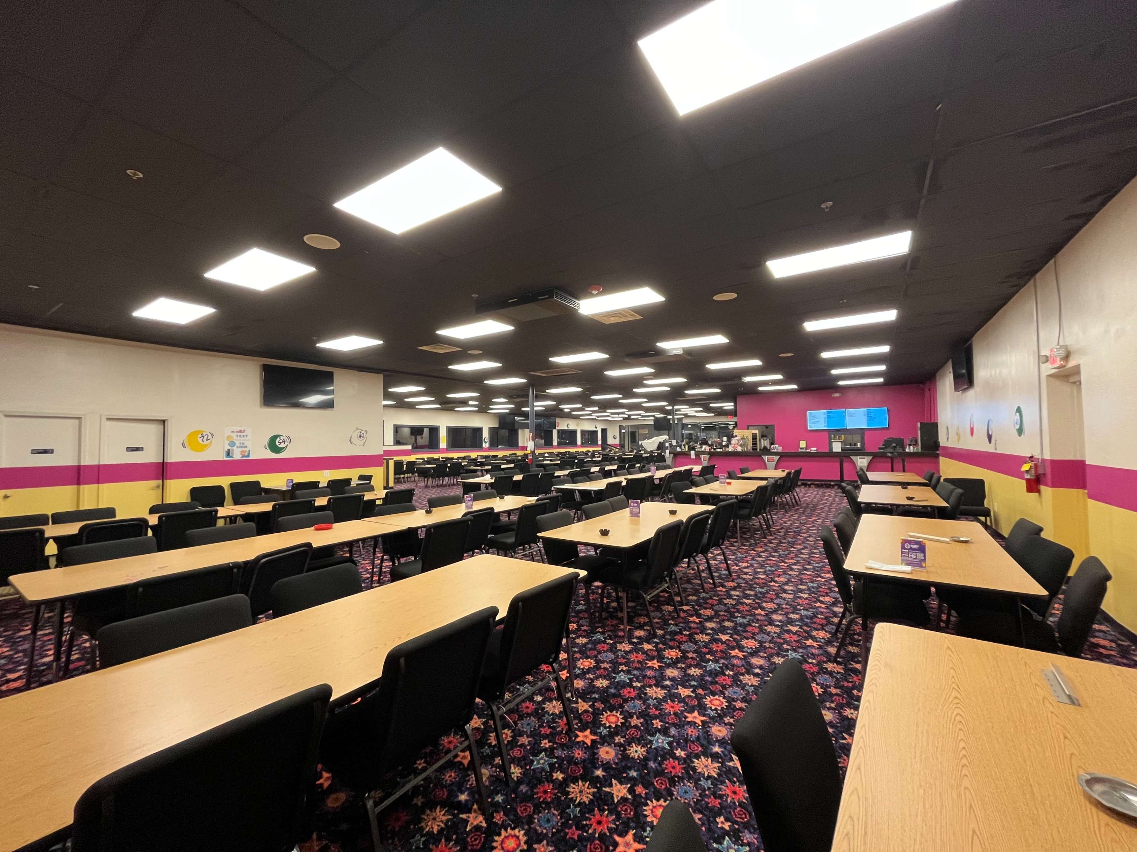 A spacious dining area with many wooden tables and black chairs arranged in rows, featuring a colorful carpet and bright overhead lighting.