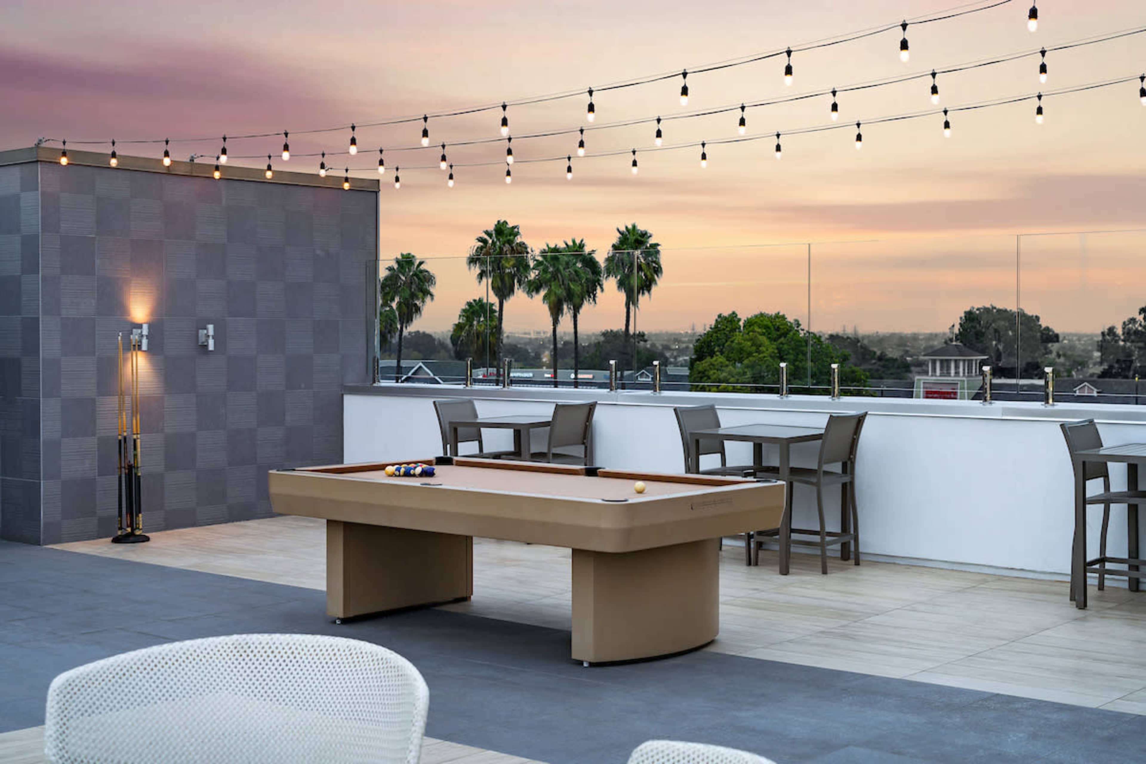 A rooftop patio with a pool table, surrounding seating, and string lights against a sunset backdrop with palm trees.