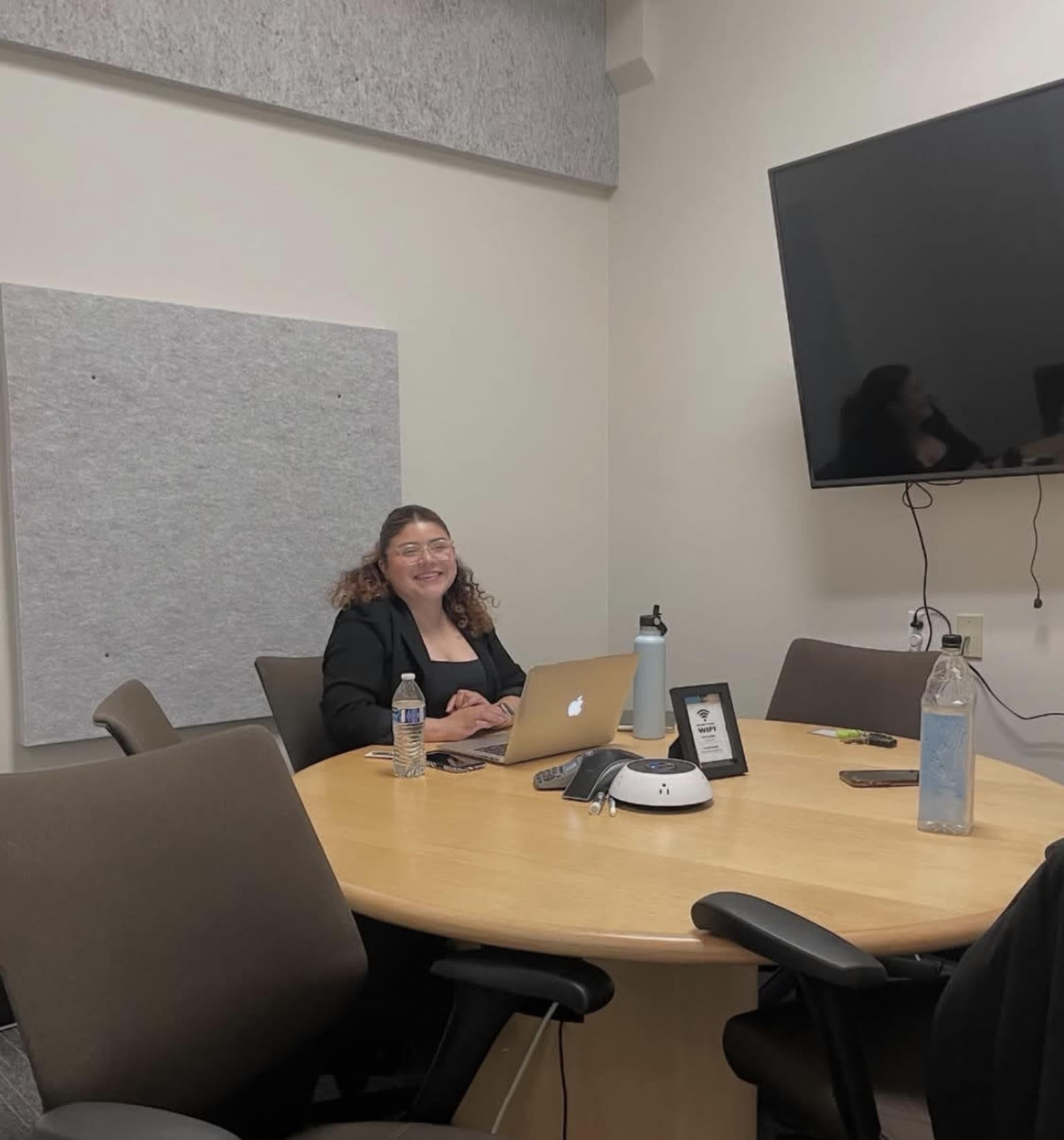 A woman sits at a conference table, working on a laptop in a meeting room with a large screen on the wall.