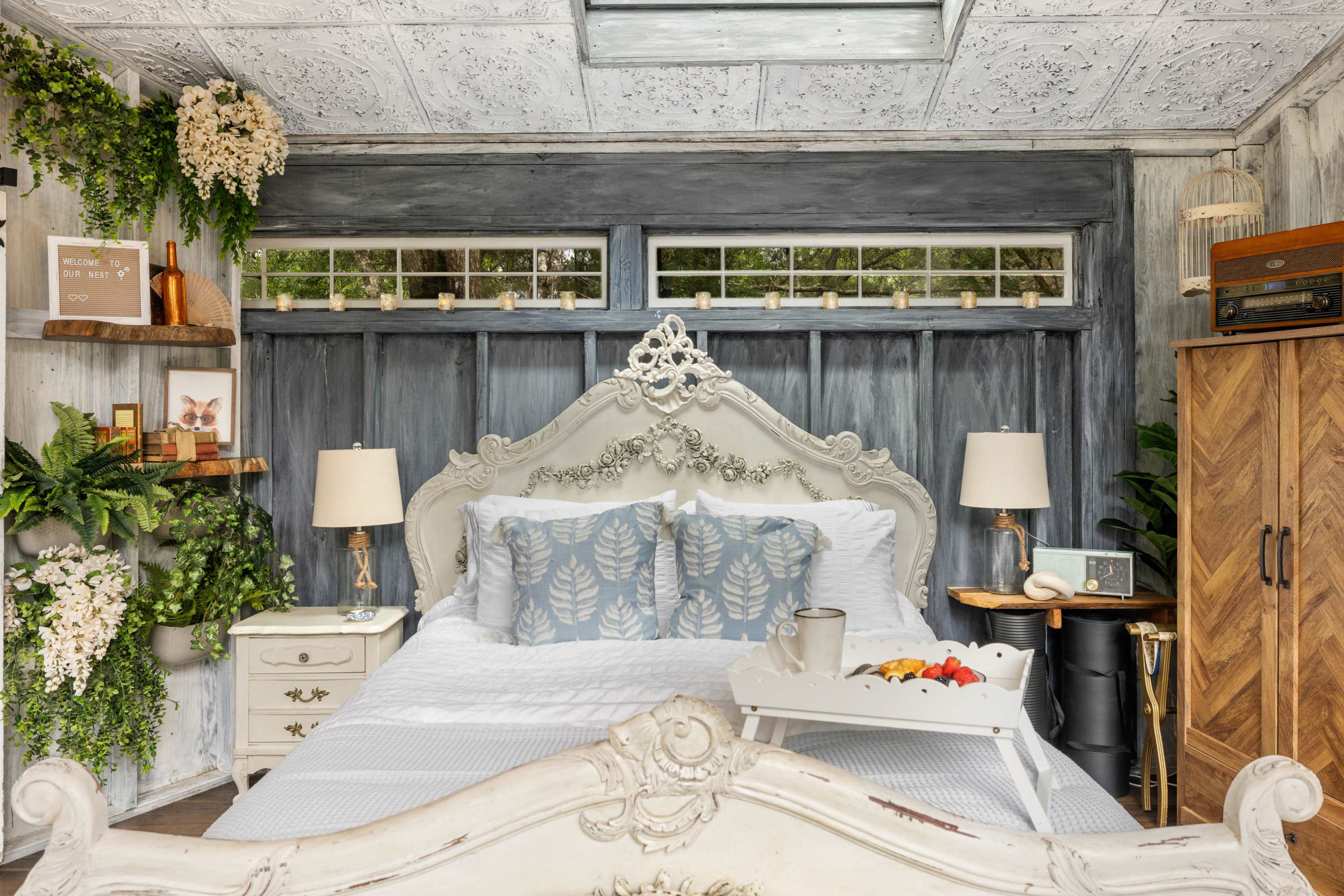 A cozy bedroom with a white bed adorned with decorative pillows, a bedside tray with fruit, and plants on the walls and shelves.