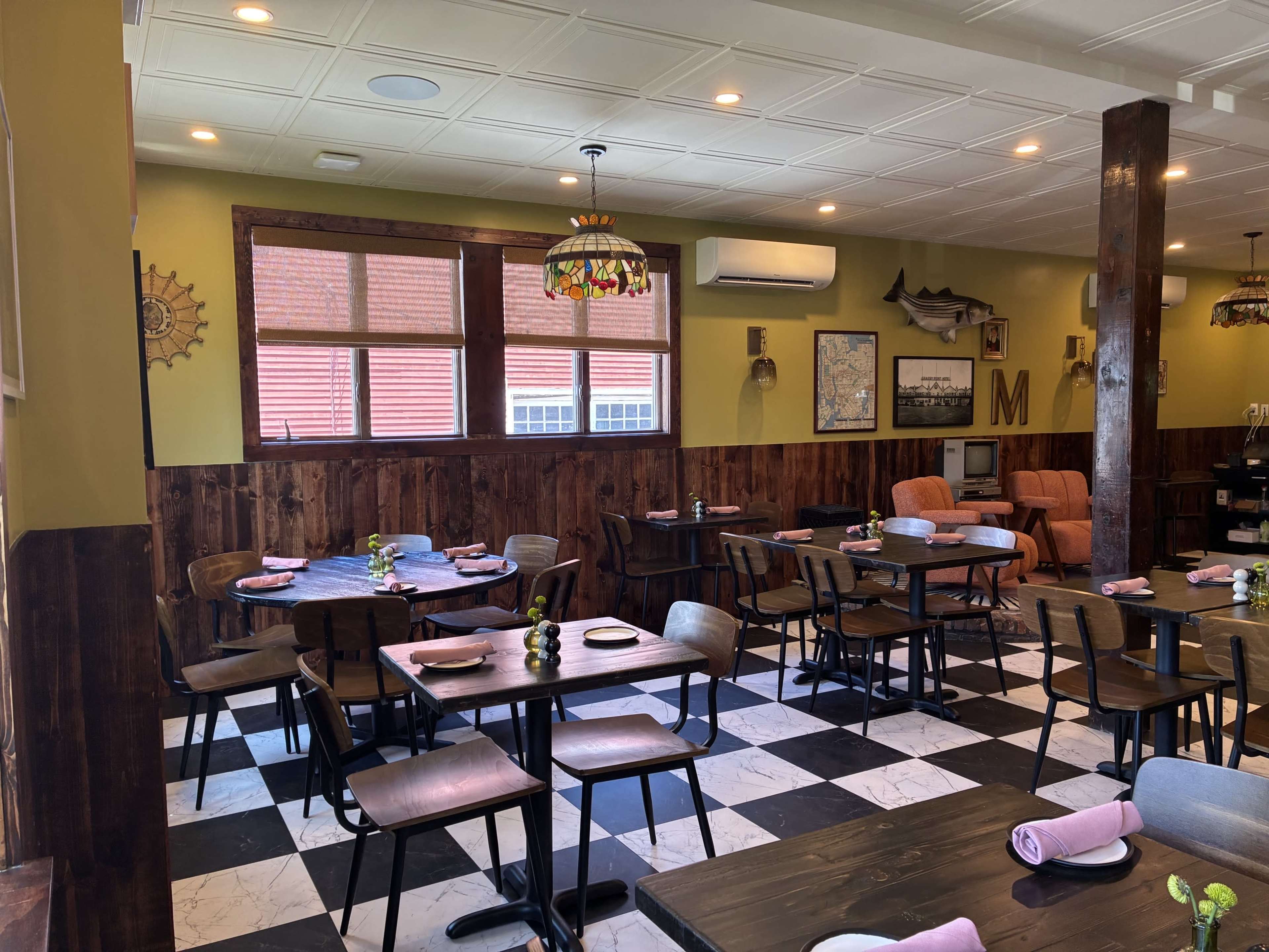 The image shows a cozy restaurant interior featuring wooden tables and chairs, patterned flooring, and a few large windows with natural light coming in.