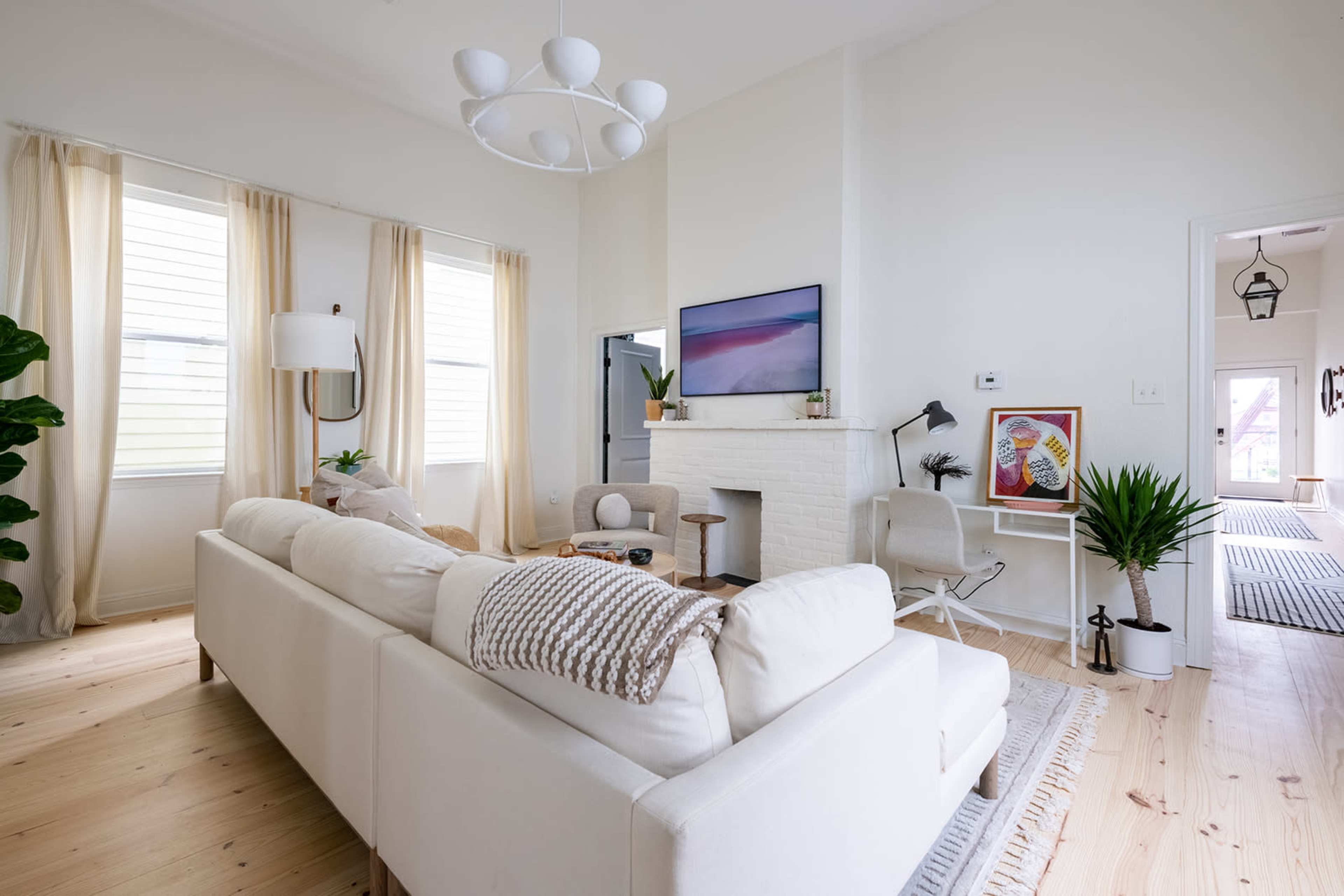 A bright living room featuring a white sofa, a modern light fixture, a fireplace, and large windows with sheer curtains.