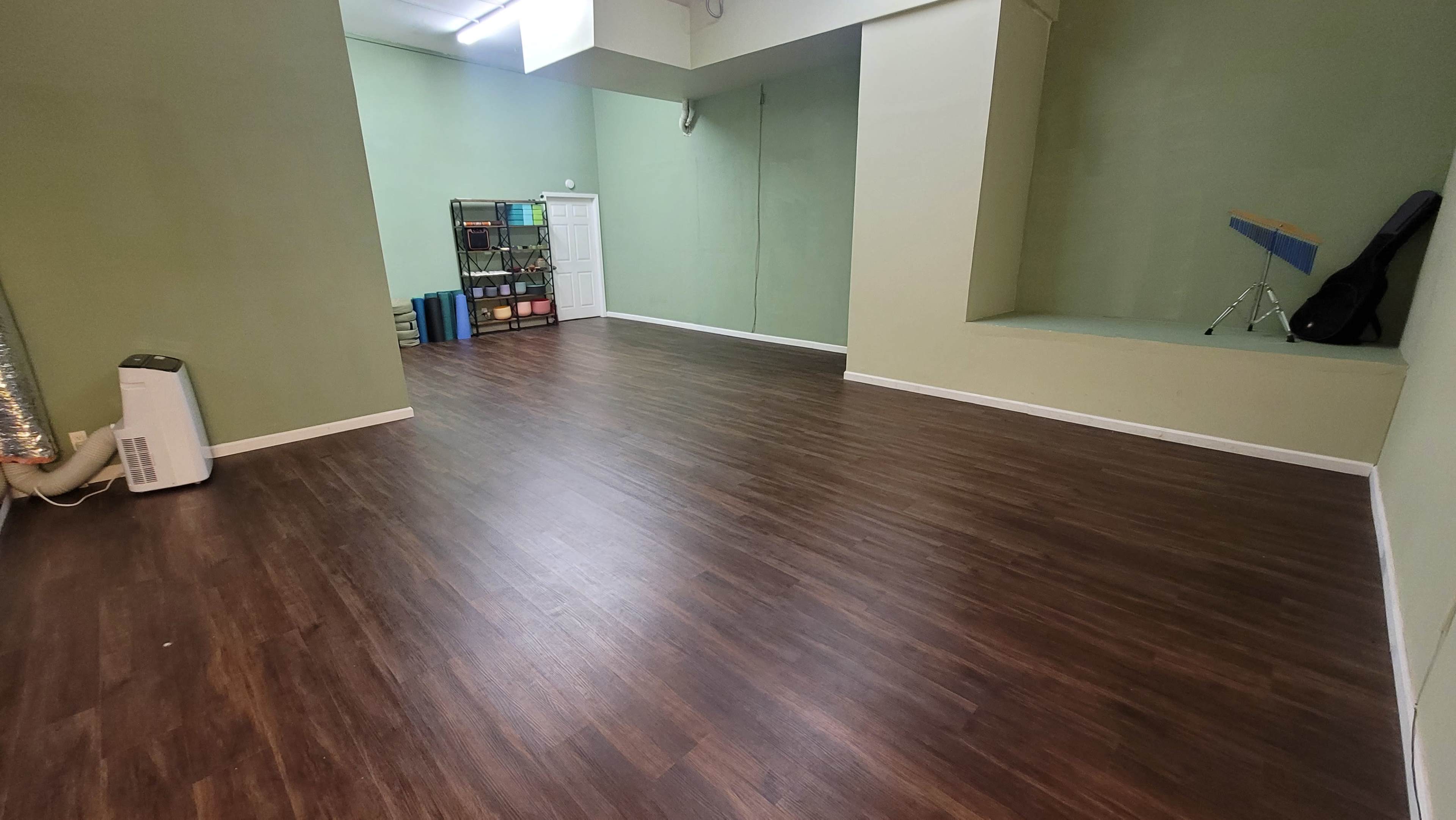 The image shows a spacious, empty room with green walls, a wooden floor, and a small stage area at one end.